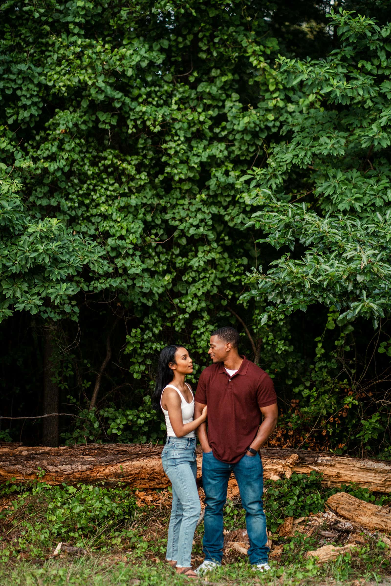raleigh engagement photographer - kayla & deandre A couple stands in front of a dense green forest. The woman, wearing a white top and jeans, faces the man in a burgundy shirt and jeans. They look at each other with a fallen tree trunk behind them.