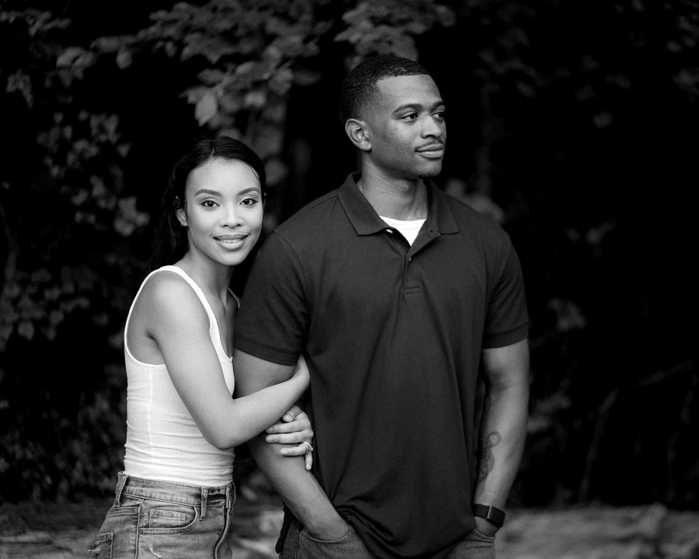 raleigh engagement photographer - kayla & deandre A black and white photo of a woman and a man standing close together outdoors. The woman, wearing a tank top and jeans, leans against the man, who is in a polo shirt and jeans. They both have a calm and content expression, with trees in the background.