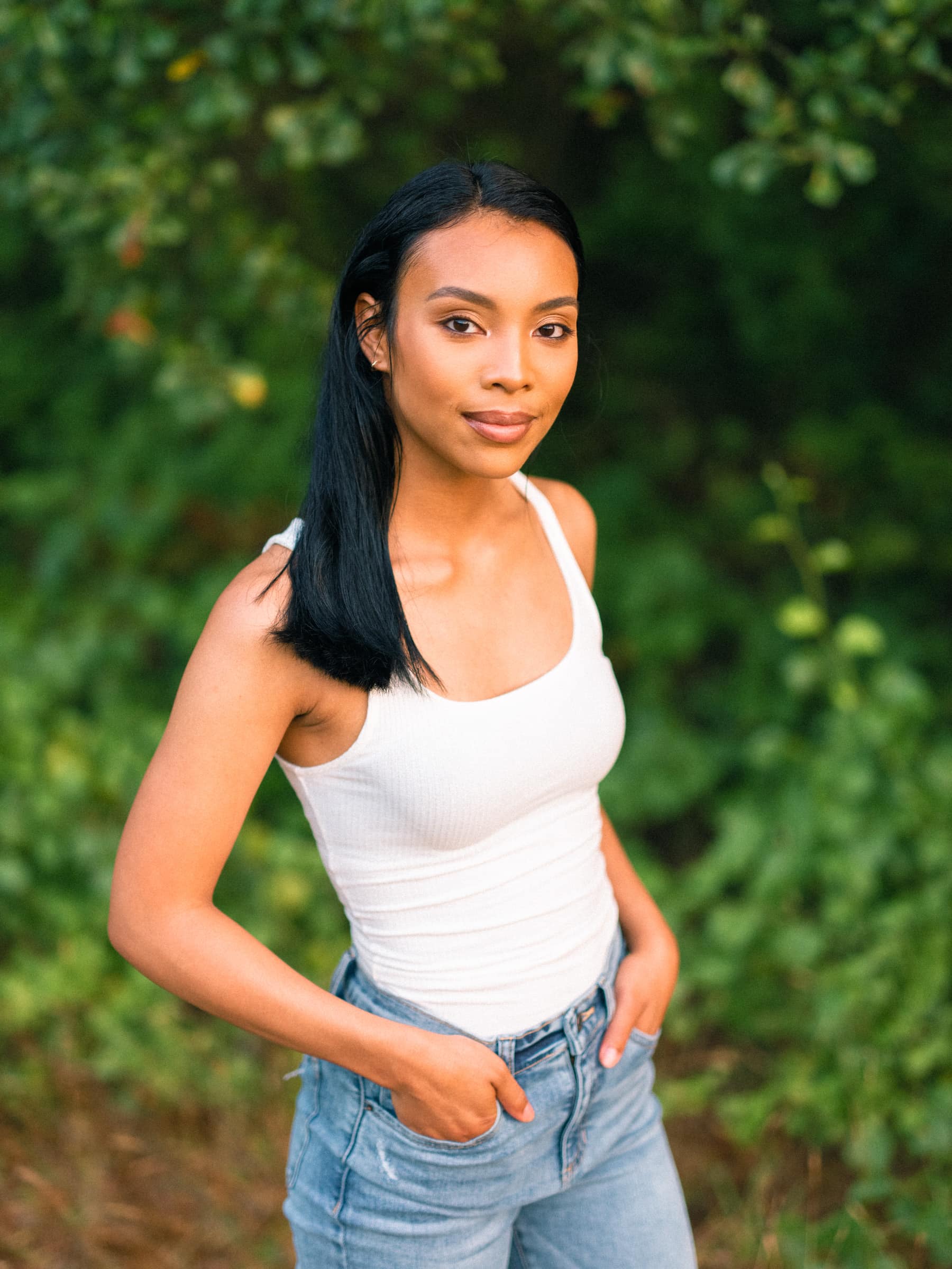 raleigh engagement photographer - kayla & deandre A woman with long black hair stands outdoors, wearing a white tank top and blue jeans. Her hands are in her pockets, and she is looking toward the camera, with green foliage in the background.