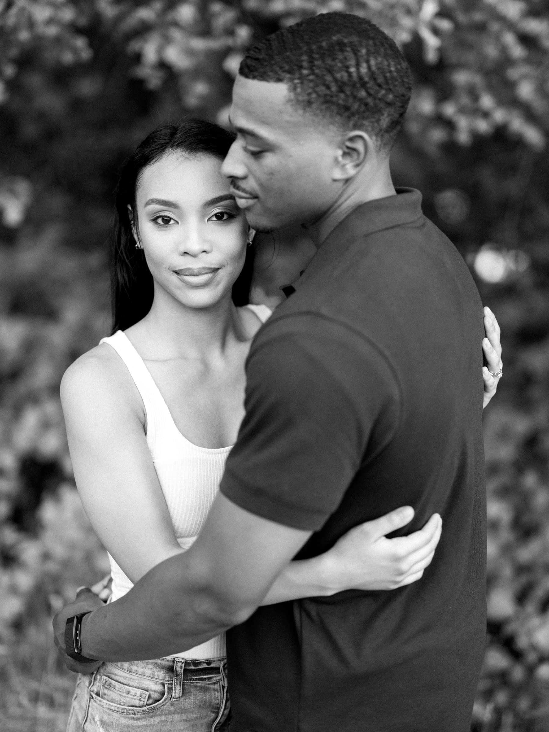 raleigh engagement photographer - kayla & deandre A black-and-white photo of a couple embracing. The woman looks into the camera with a gentle expression, wearing a sleeveless top and jeans. The man, in a short-sleeve shirt, faces her, holding her in a serene outdoor setting.