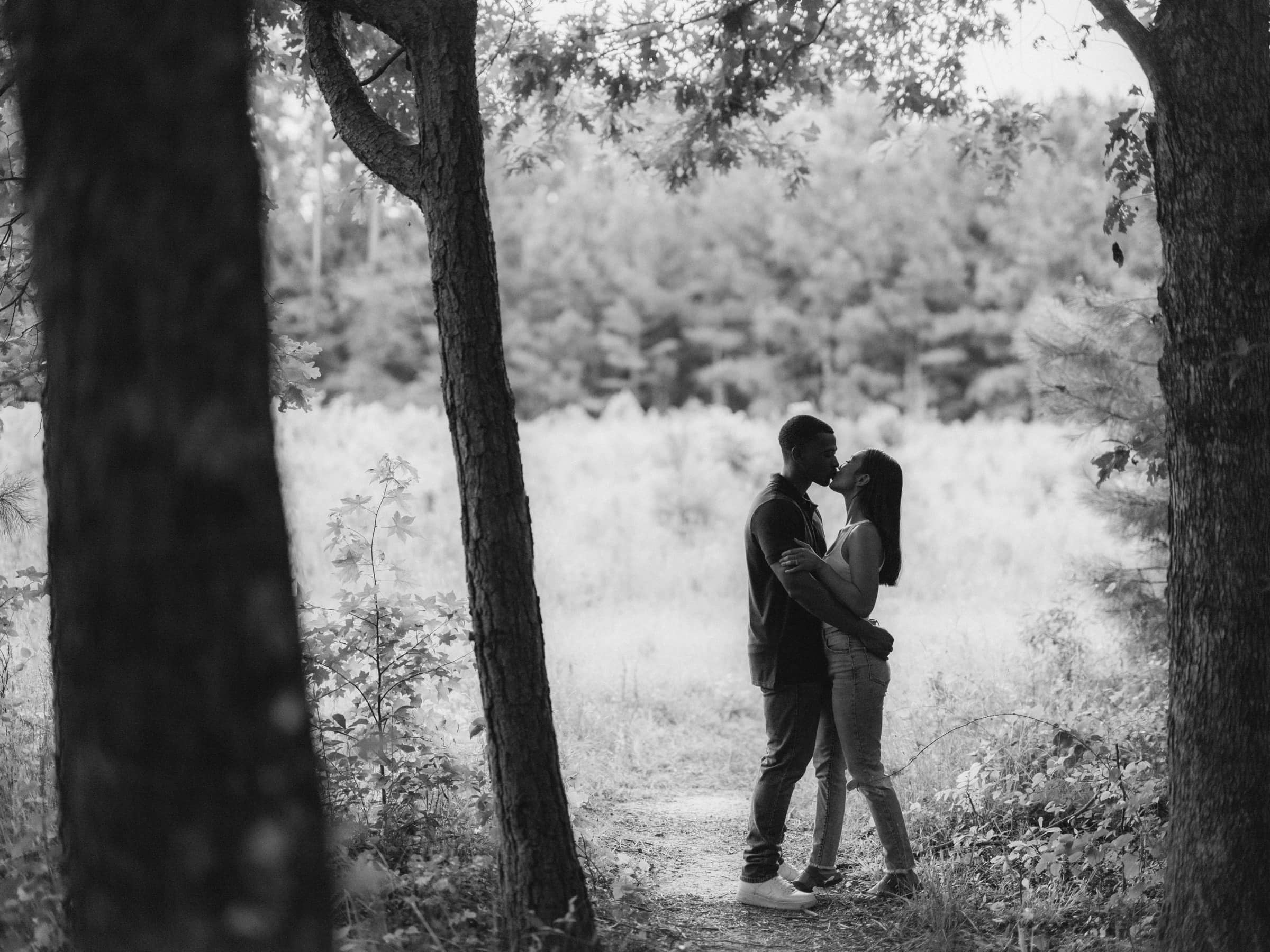 raleigh engagement photographer - kayla & deandre A couple stands closely, embracing and kissing in a forest path framed by trees. The scene is in black and white, with a serene, natural backdrop of trees and foliage.