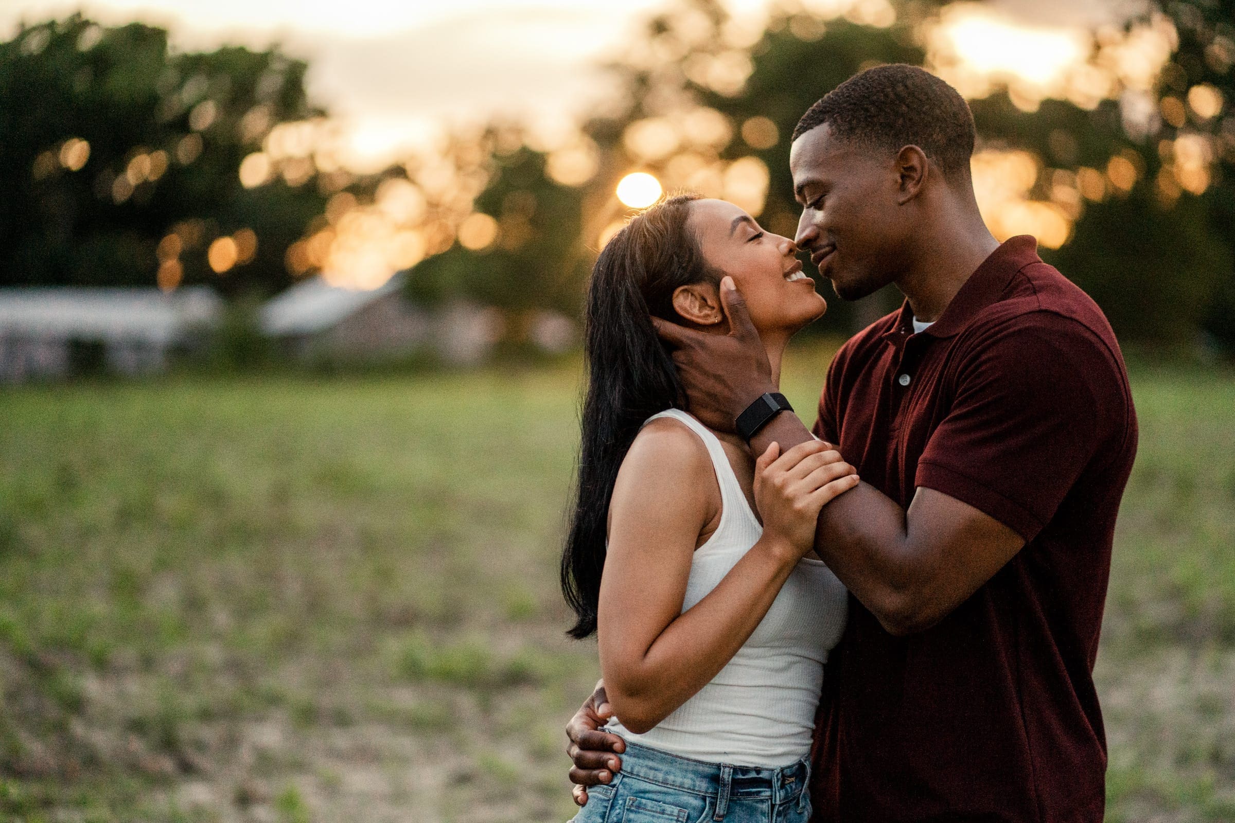 raleigh engagement photographer - kayla & deandre A couple embraces in a field at sunset. They are smiling and gazing into each others eyes, with one wearing a white tank top and the other a burgundy shirt. Trees and a soft, glowing sky are in the background.