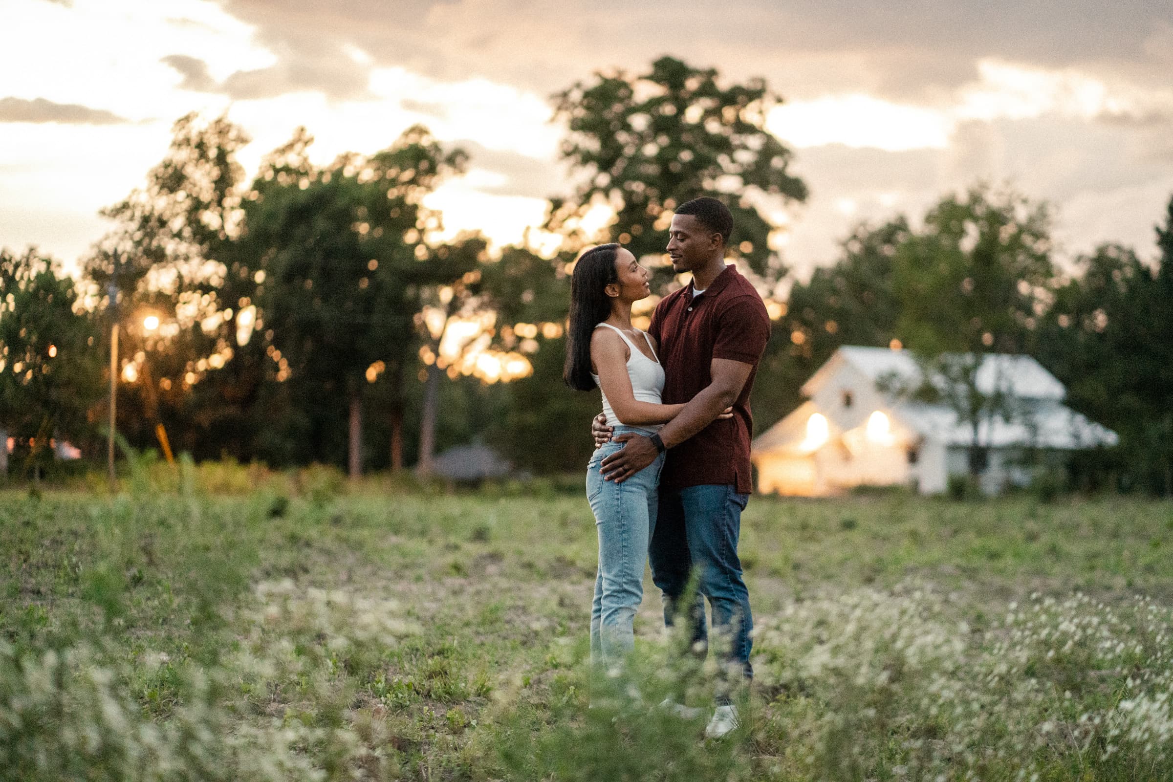 raleigh engagement photographer - kayla & deandre A couple embraces in a grassy field at sunset. They stand facing each other, smiling, with trees and a house visible in the background. The sky is partially cloudy with a warm glow.