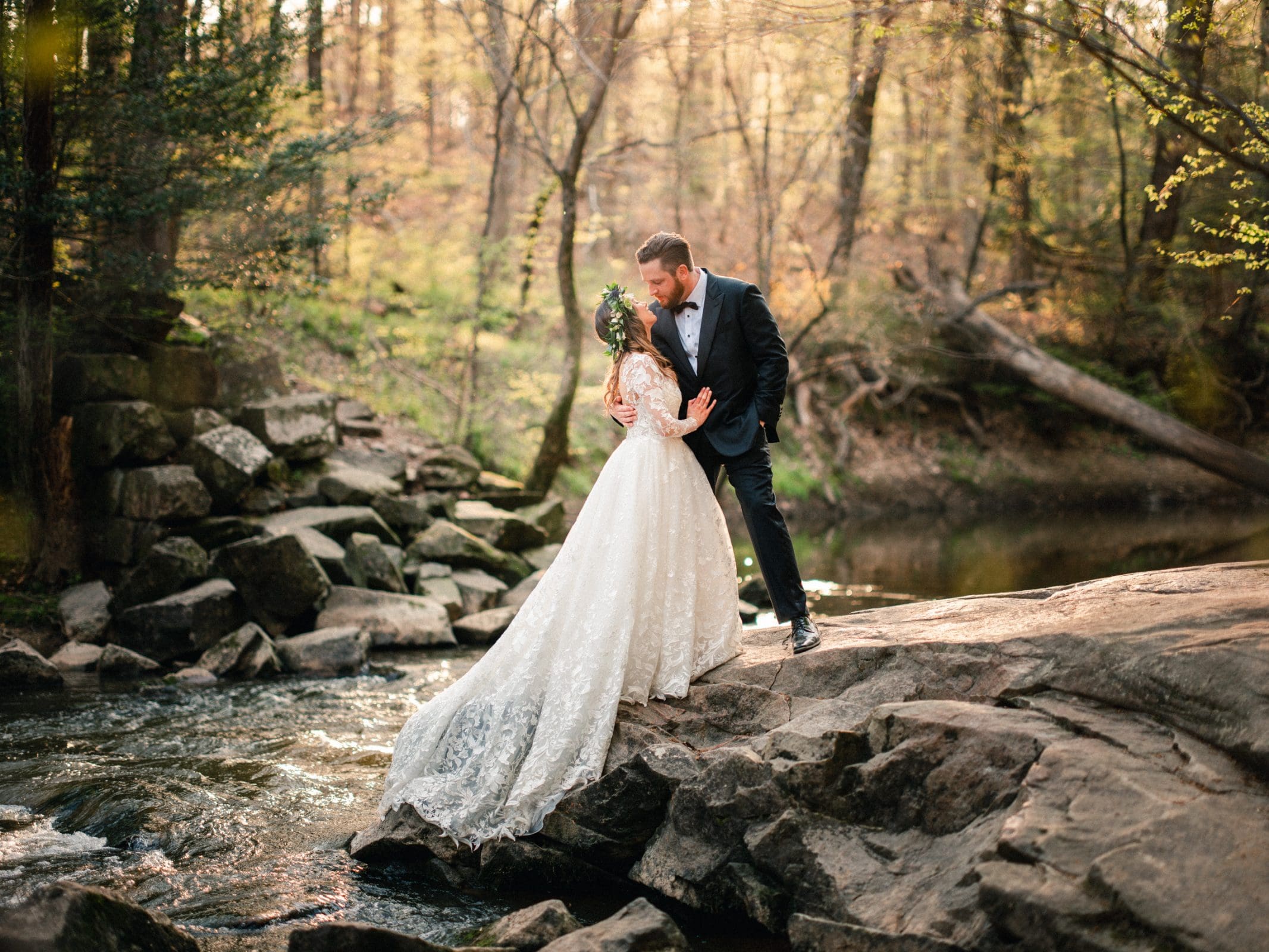 A couple in wedding attire stands on rocks by a serene forest stream. Captured by Raleigh NC wedding photographers, the groom, in a navy suit, gently kisses the bride, who wears a flowing white gown and floral headpiece. Sunlight filters through the trees, creating a warm, romantic atmosphere.
