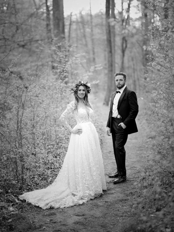 A bride and groom stand on a forest path, captured by Raleigh NC wedding photographers. The bride wears a long lace gown and floral crown, while the groom is dressed in a dark suit and bow tie. The trees create a natural backdrop for the couple in this stunning black-and-white image.