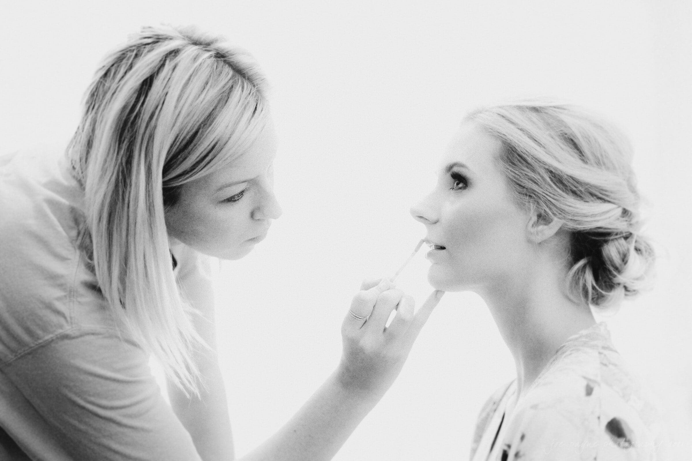 A makeup artist applies lipstick to a woman with styled hair in an elegant updo, both focused and set against a bright, softly lit background. The image is in black and white, giving it a timeless feel.