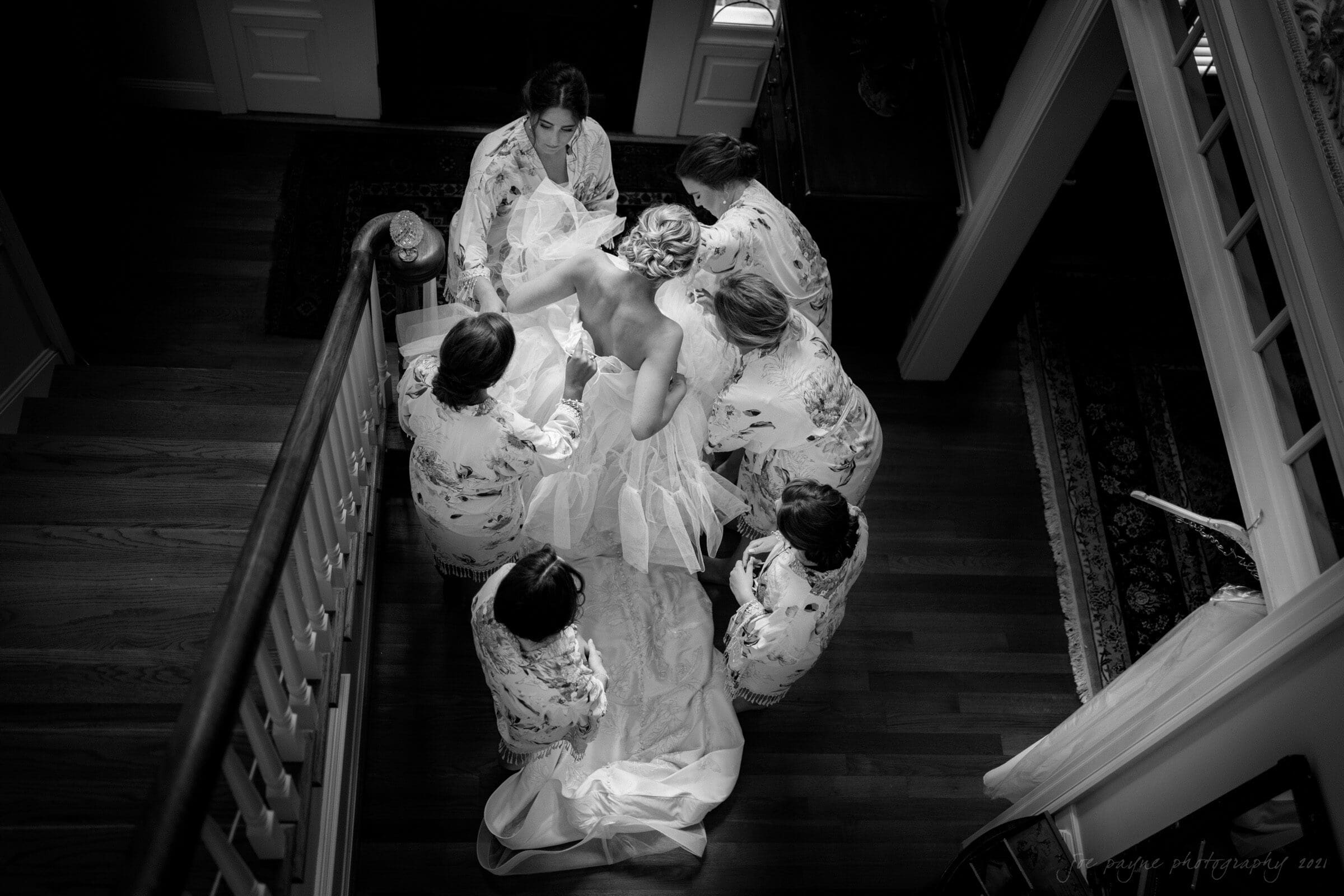 A black and white photo shows six people in floral robes surrounding a bride on a wooden staircase. They are adjusting her dress train. Light streams in from windows, creating a serene and intimate atmosphere.