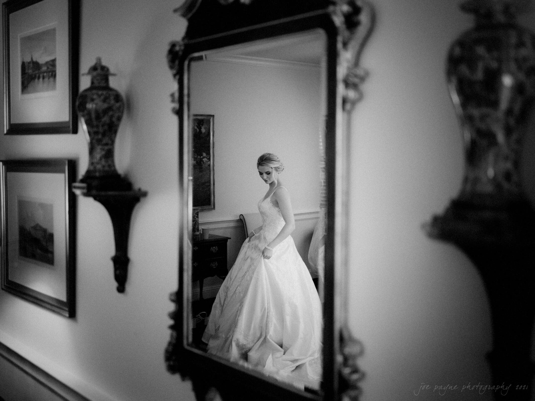 A black and white photo shows a bride in a flowing gown, reflected in an ornate mirror. She stands gracefully in a softly lit room, surrounded by framed pictures and decorative vases.