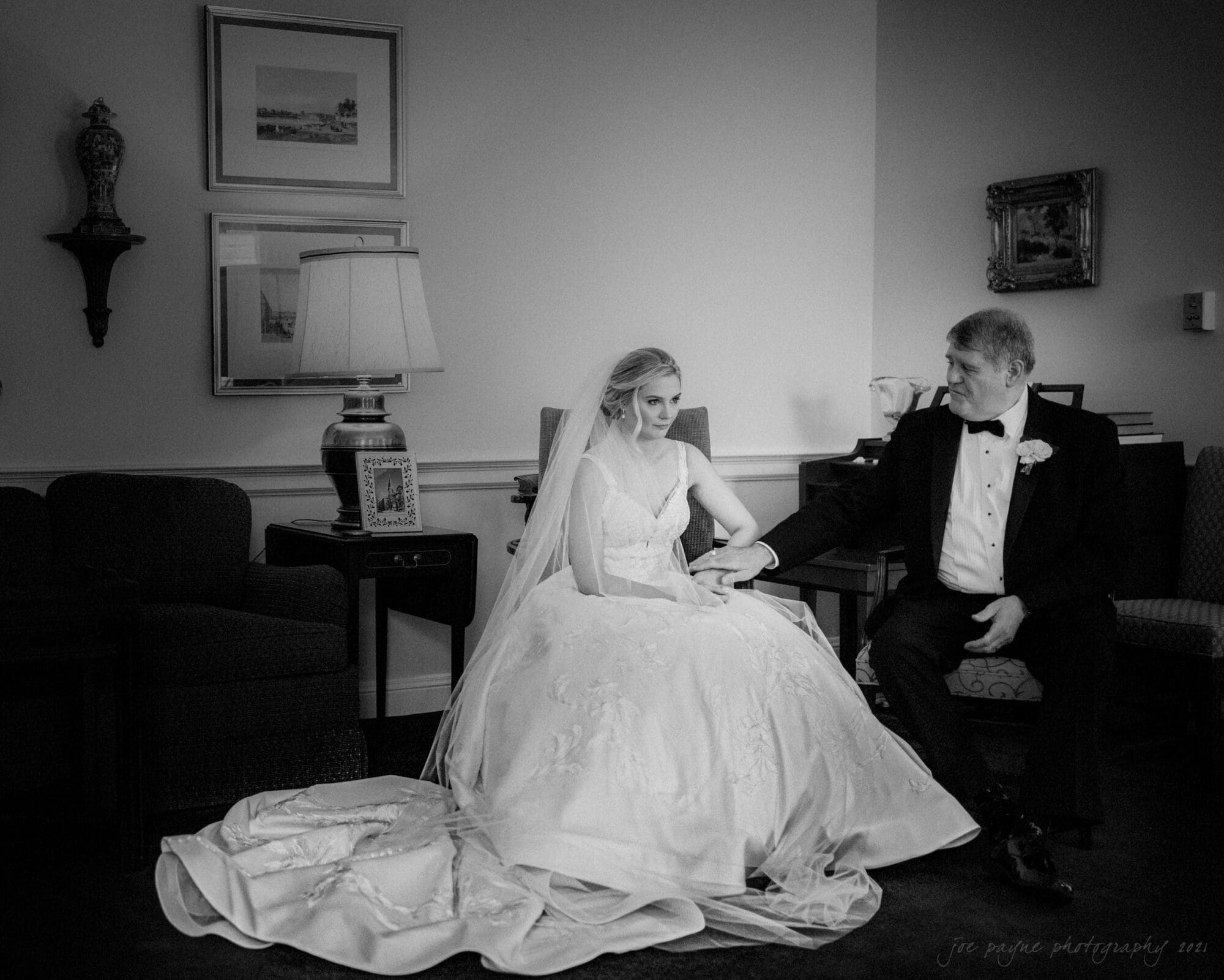 A bride in a wedding gown and veil sits beside an older man in a tuxedo. They appear to share a moment together in a dimly lit room adorned with framed art and a lamp. The atmosphere is serene and intimate.