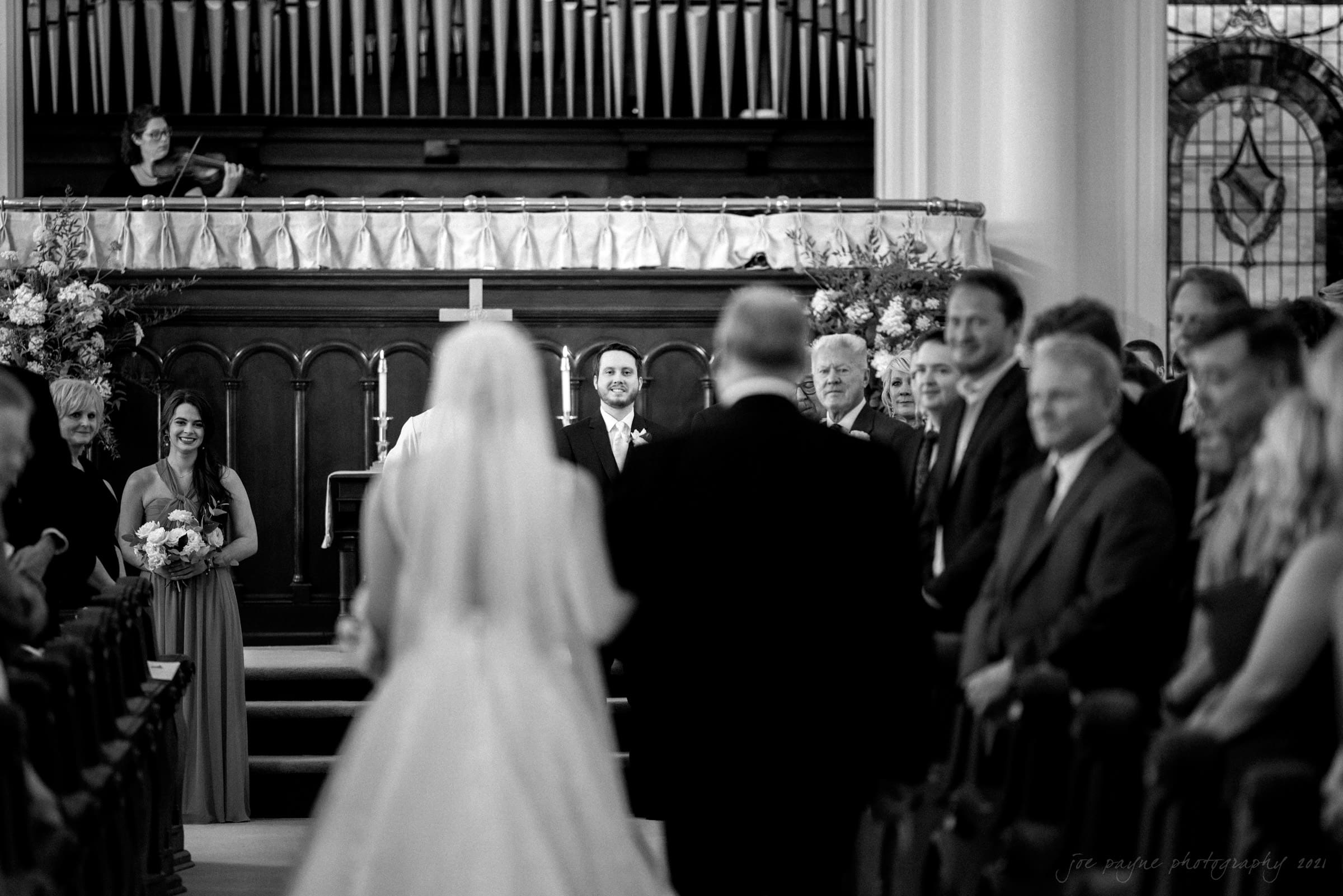 A black and white photo of a bride walking down the aisle with her father in a church. Guests stand on either side, and the groom waits at the altar. A musician plays a violin on the balcony above.
