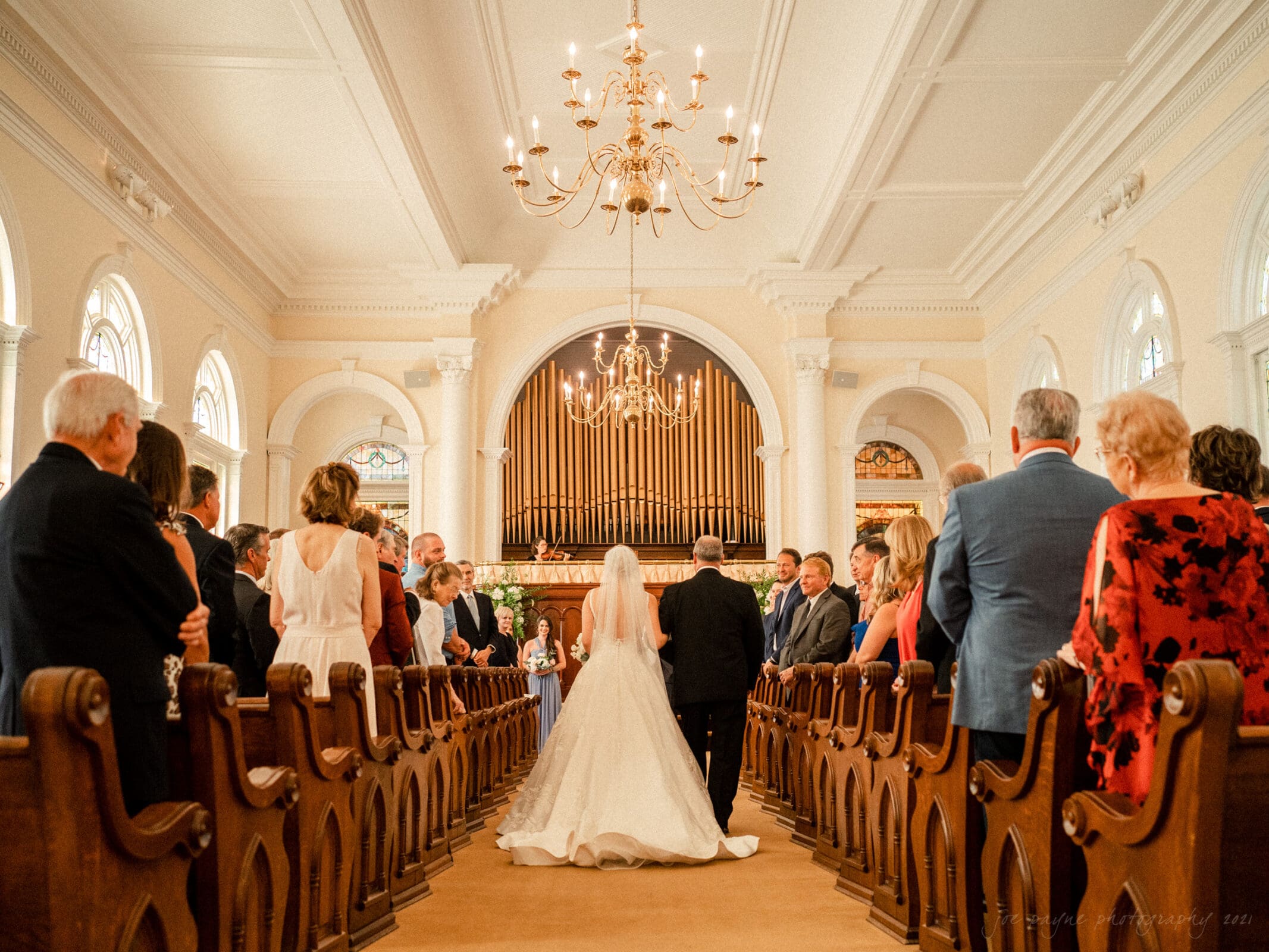 A bride in a white gown walks down the aisle of a grand church, accompanied by a man in a suit, towards a group waiting at the altar. The church is adorned with chandeliers and large organ pipes, and the pews are filled with well-dressed guests.