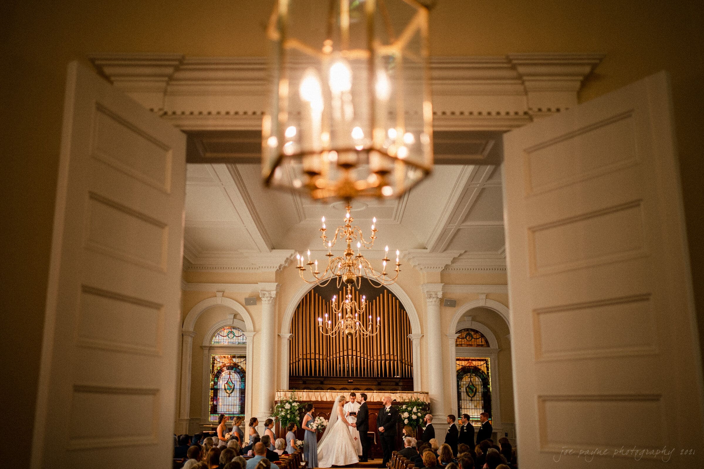 A bride and groom stand at the altar in a church, surrounded by their bridal party. The interior features a large organ, stained glass windows, and a chandelier. Guests are seated, with the scene viewed from an open door in the foreground.