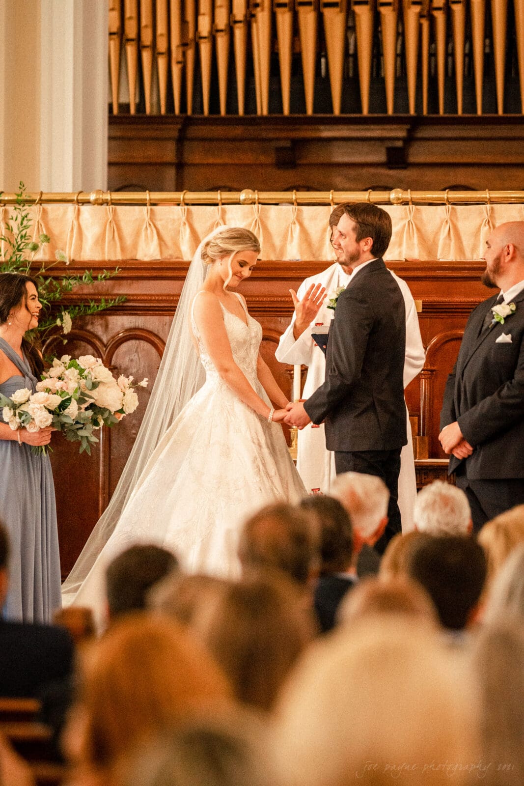 A bride and groom stand at the altar in a church, holding hands during their wedding ceremony. The bride wears a white gown and veil, while the groom is in a dark suit. Bridesmaids and groomsmen are seen nearby, with guests seated in the foreground.