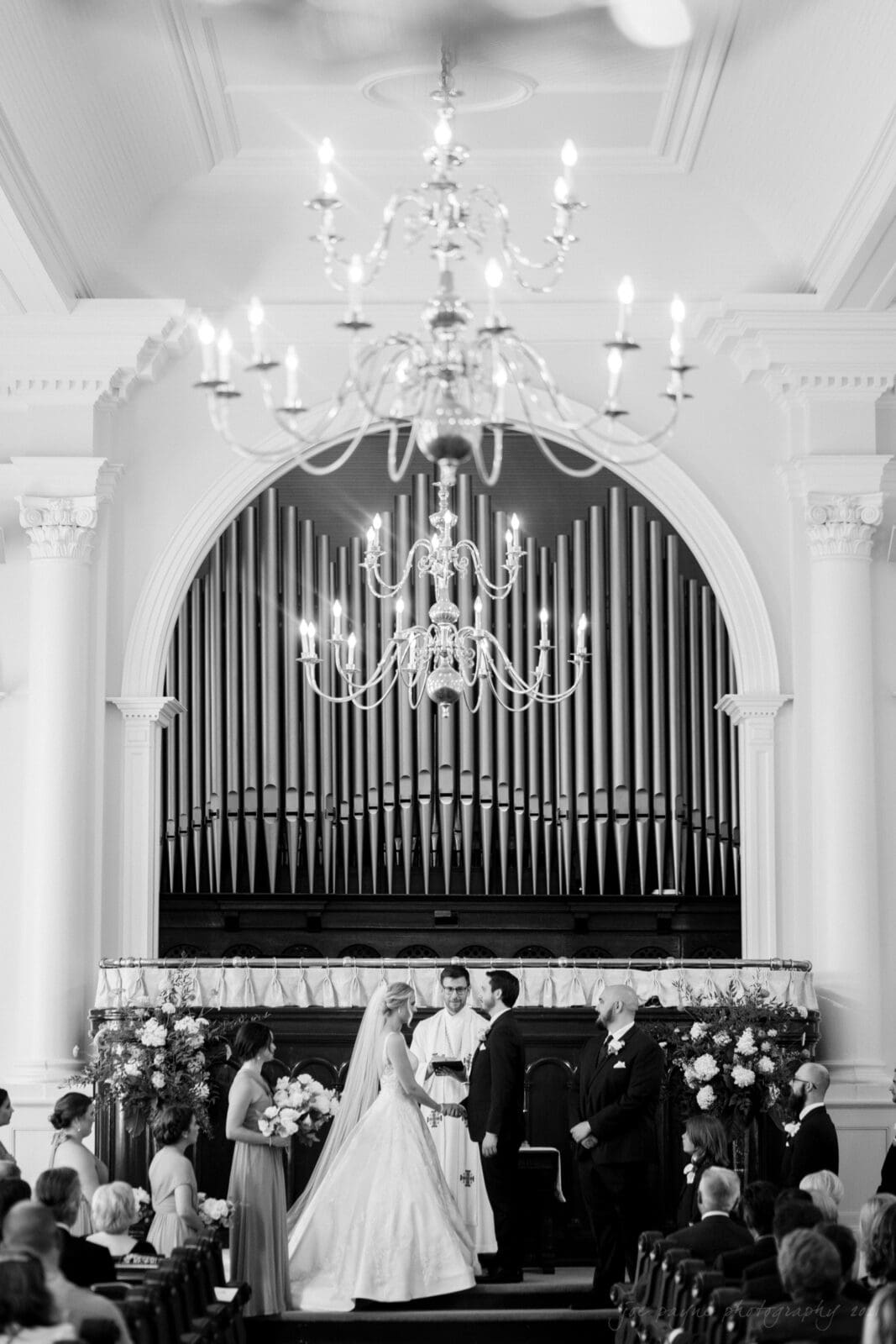 A black and white photo of a couple getting married in a church. The bride and groom stand at the altar with a priest. A large chandelier hangs above, and an organ pipe structure is in the background. Guests are seated on either side.