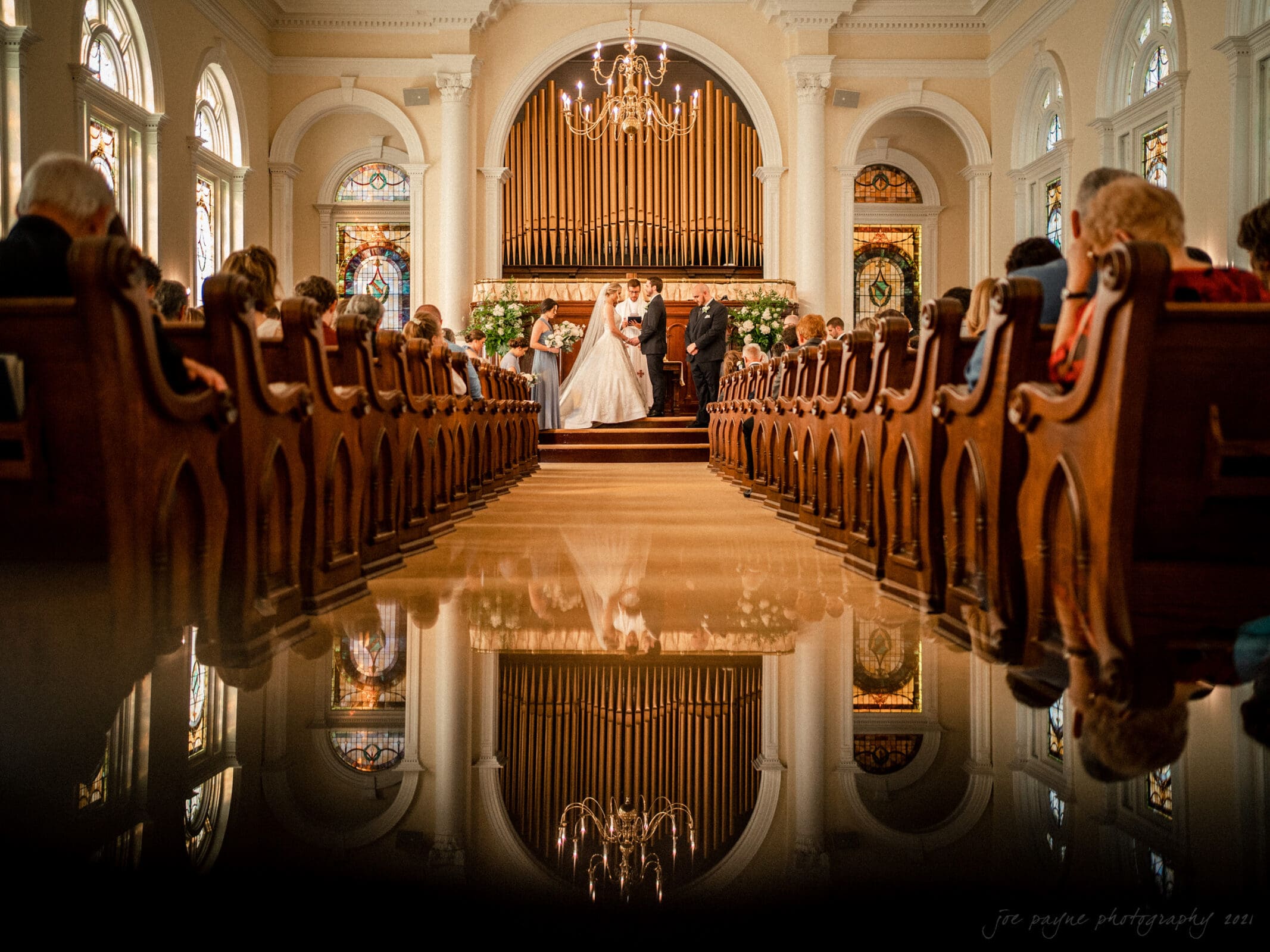 A bride and groom stand at the altar in a church during a wedding ceremony. Guests are seated in wooden pews on either side. The reflection of the scene is visible on a shiny floor. Stained glass windows and a large organ are in the background.