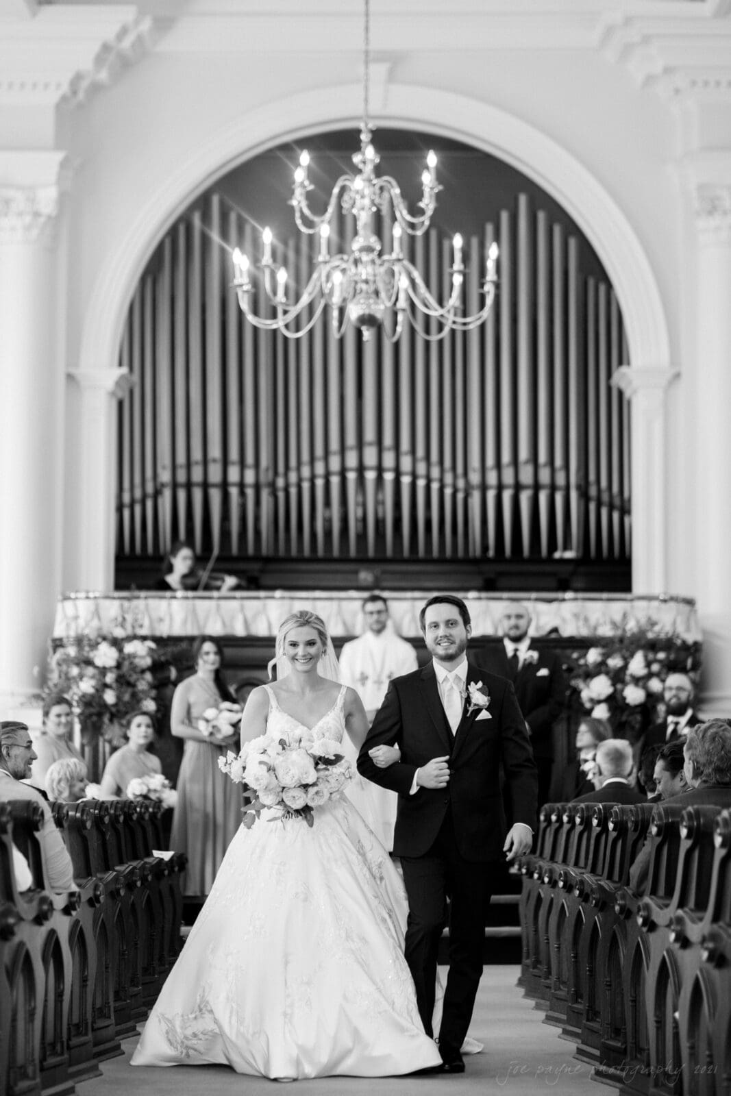 A bride and groom walk arm in arm down the aisle of a church. The bride wears a flowing white gown and holds a bouquet, while the groom is in a dark suit. Guests are seated on either side, and a large organ is visible in the background.