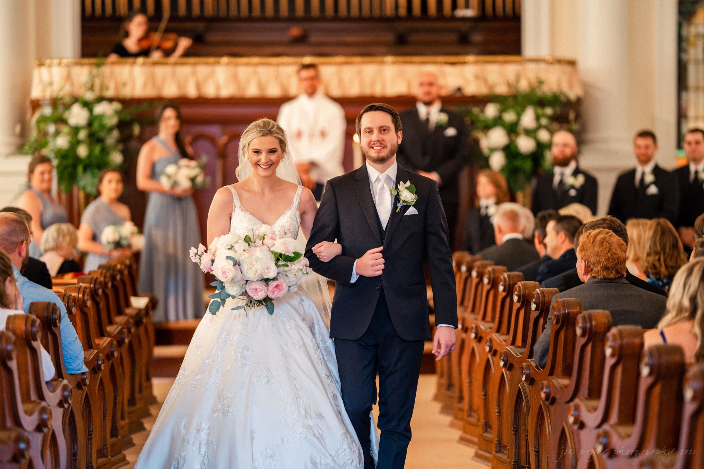 A bride and groom walk down the aisle in a church, surrounded by seated guests. The bride is holding a bouquet of white and pink flowers, and the groom is in a dark suit. Floral arrangements adorn the background.