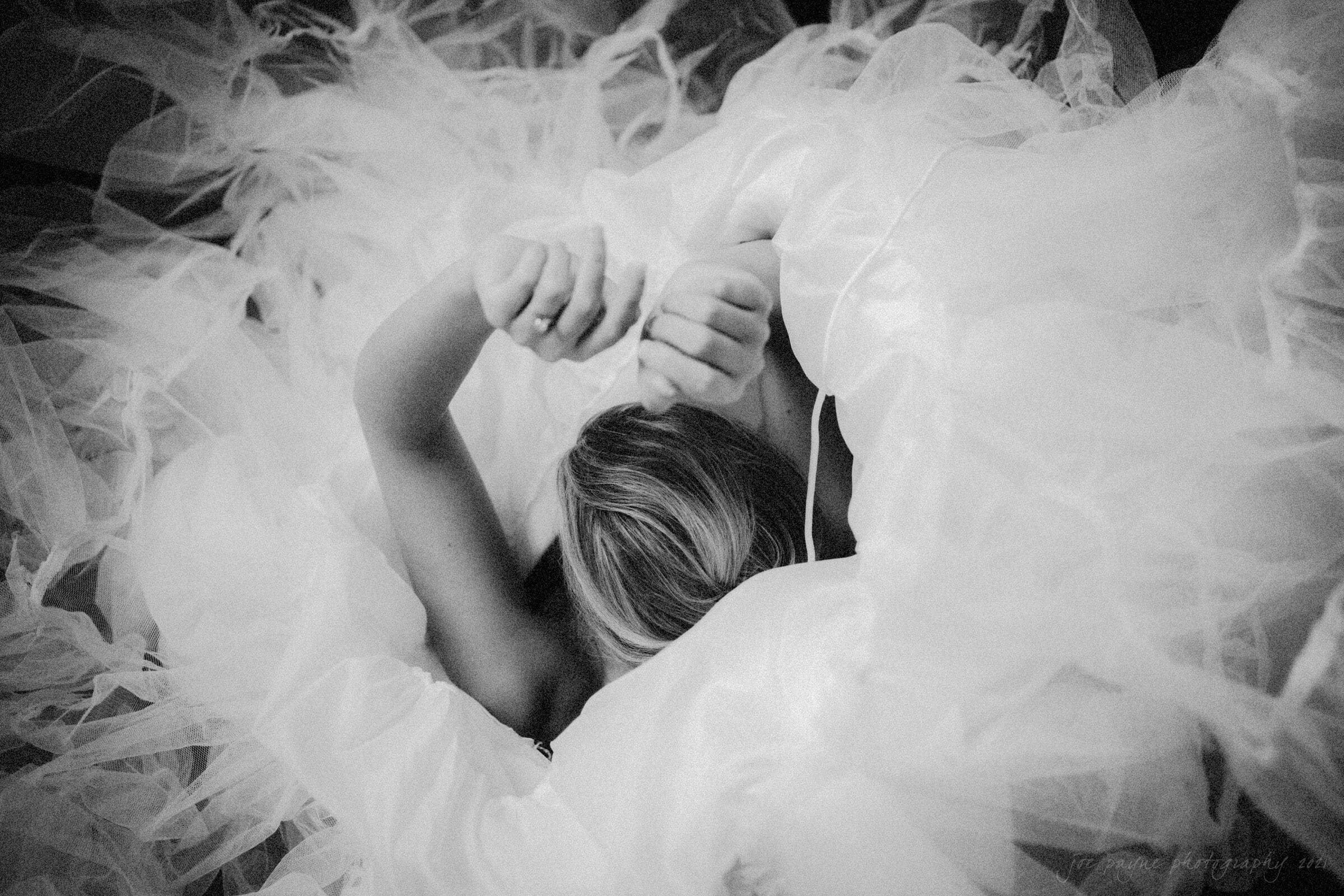 A person is lying down, partially covered by a large, swirling mass of soft, white fabric, with their hands gently resting above their head. The scene is ethereal and dreamlike, captured in black and white.