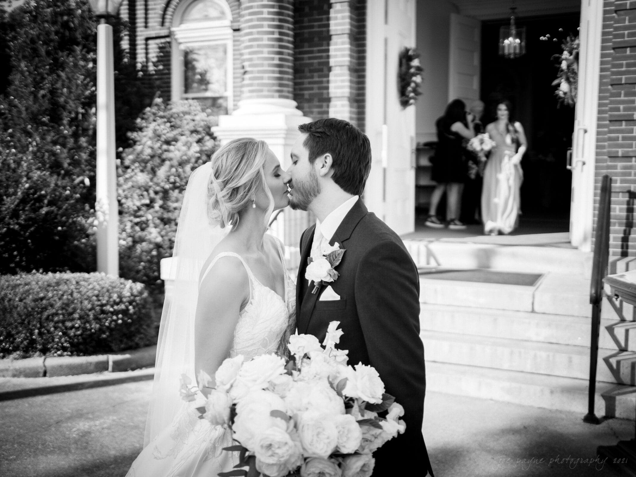 A newlywed couple shares a kiss outside a church. The bride holds a bouquet of flowers and wears a wedding dress with a veil, while the groom wears a suit. Bridesmaids are visible in the background near the church entrance.