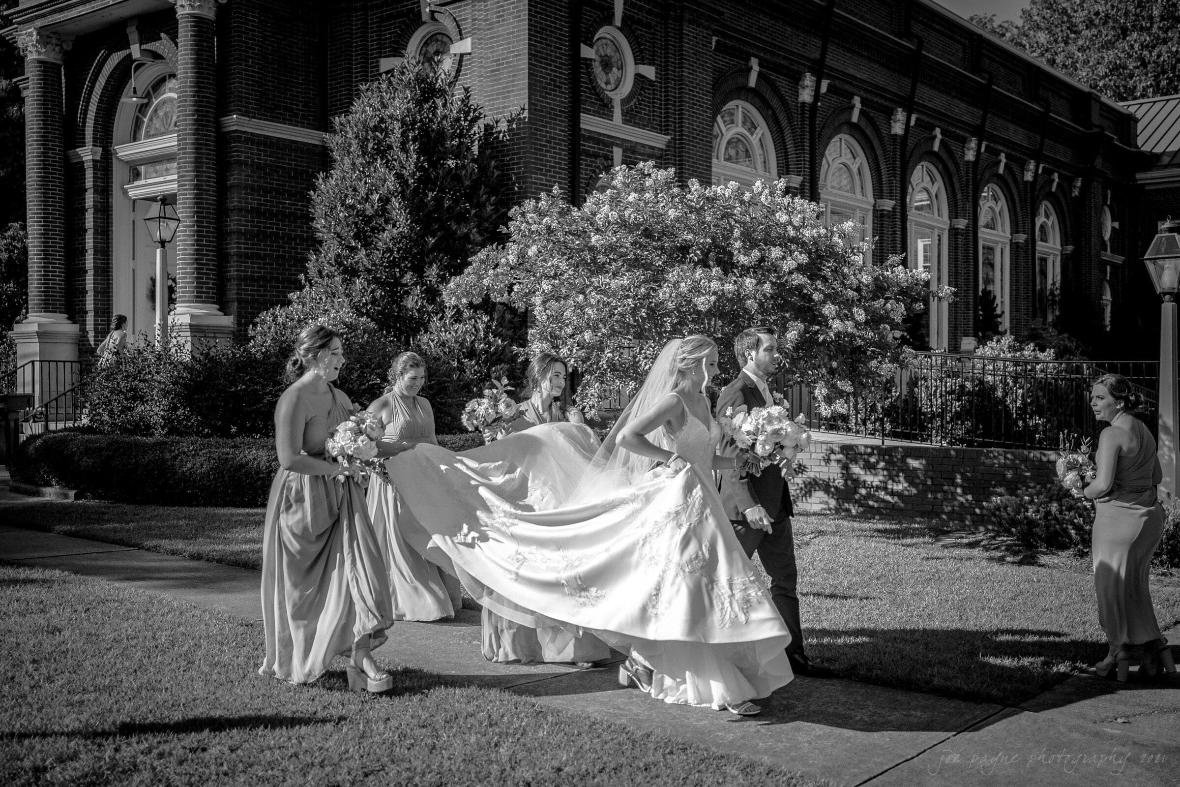 A bride in a flowing dress walks with a groom beside a historic brick building. Bridesmaids in elegant dresses hold her train. They are surrounded by lush greenery and the scene is sunlit, highlighting the joyful occasion.