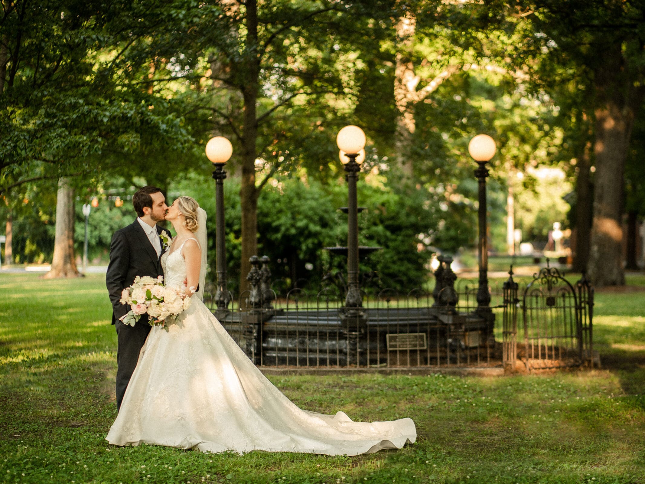 A bride and groom share a kiss in a lush, green park. The bride wears a flowing white gown, holding a bouquet of white and pink flowers. Behind them is an ornate black fountain adorned with spherical lights, surrounded by trees.