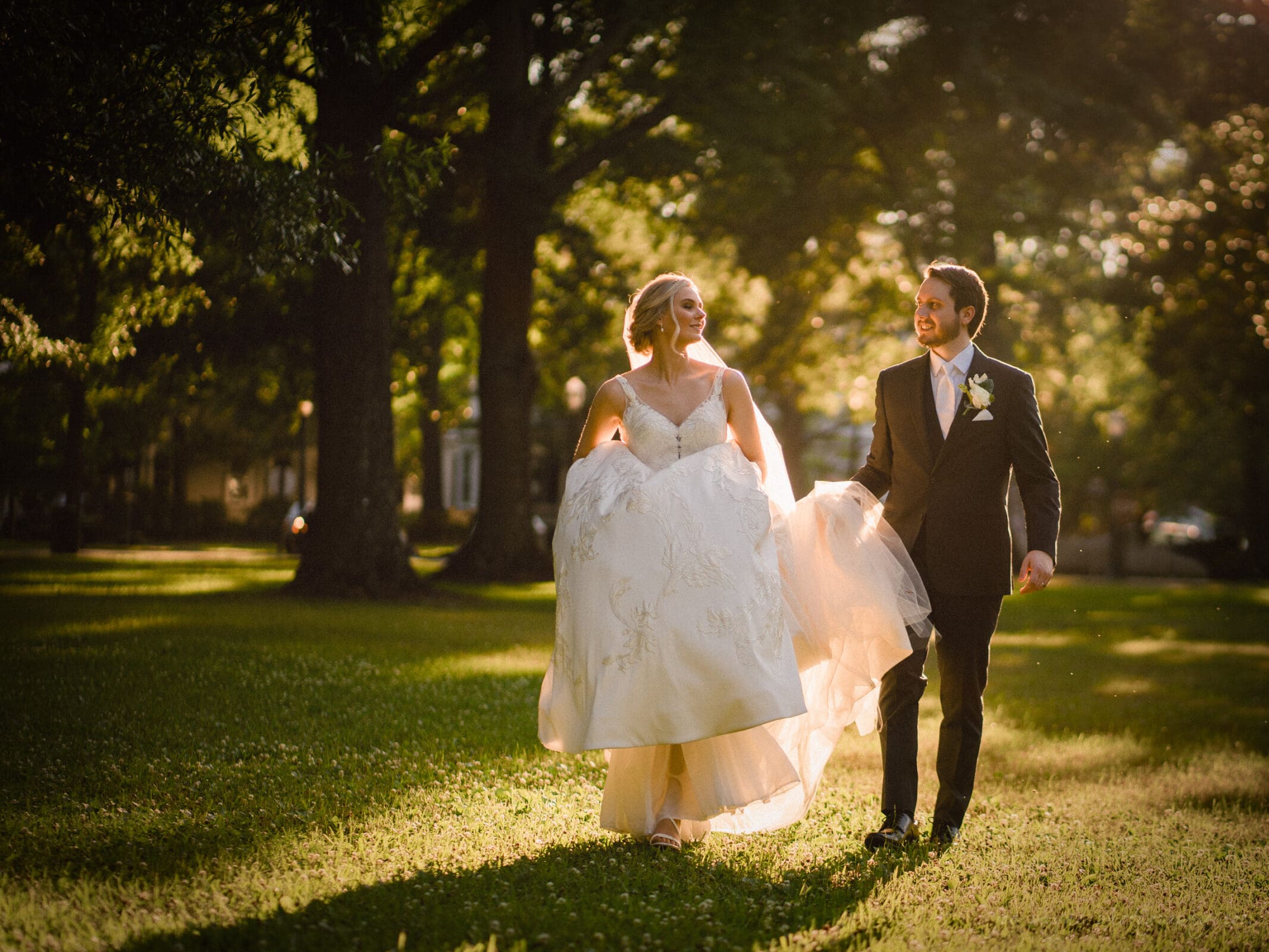 A bride and groom walk hand in hand through a sunlit, grassy park. The bride holds up her white dress, and both are smiling at each other, surrounded by trees and soft, golden light.