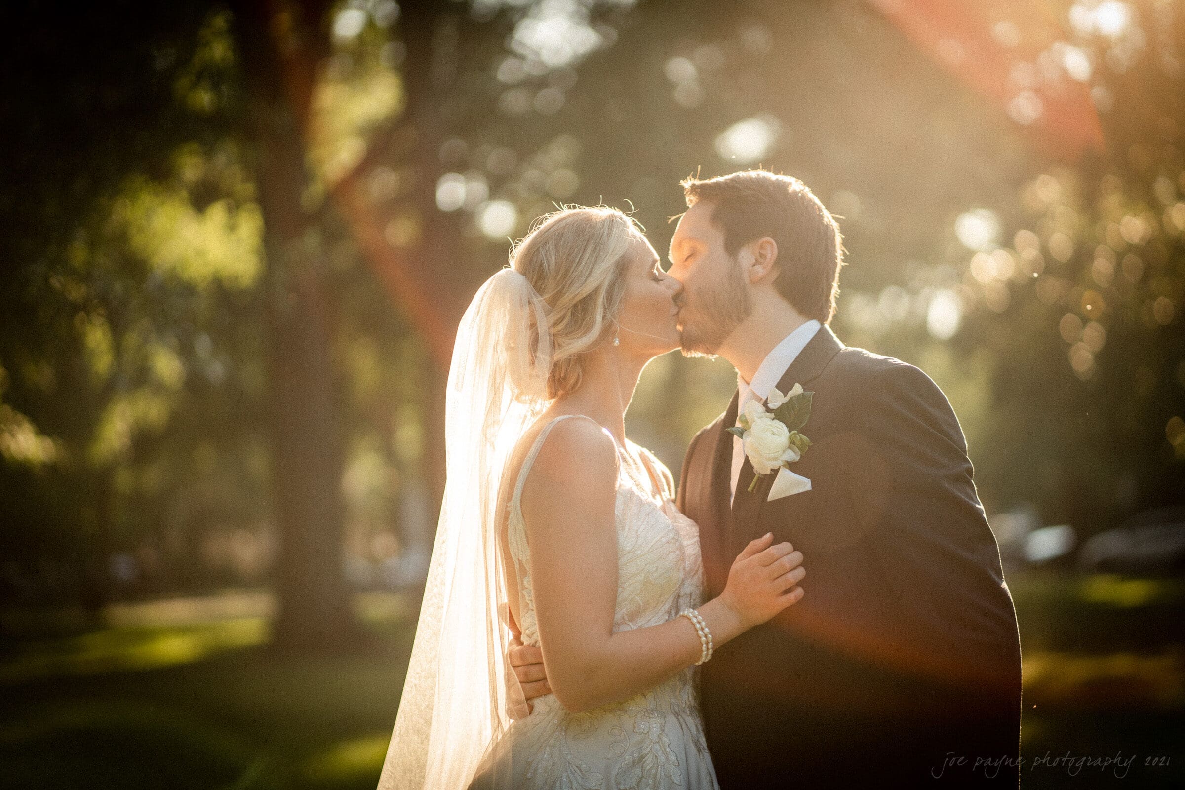 A bride and groom share a kiss outdoors in a sunlit, grassy area. The bride wears a white gown and veil, while the groom is in a dark suit with a white boutonniere. Sunlight filters through the trees, creating a warm, romantic atmosphere.
