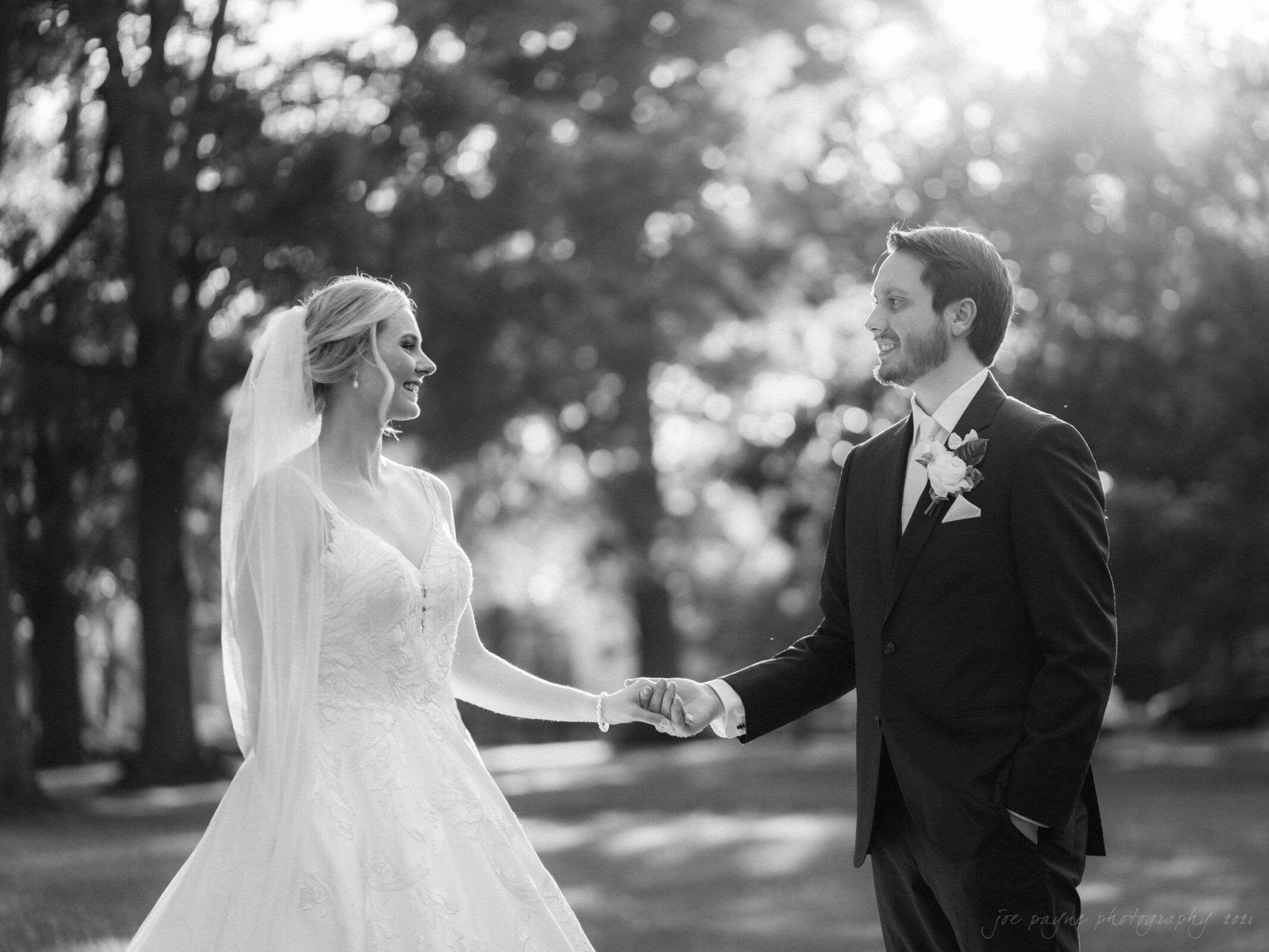 A bride and groom hold hands and smile at each other in a sunlit park. The bride wears a flowing gown and veil, while the groom is in a suit with a boutonniere. Trees and soft sunlight create a serene backdrop.