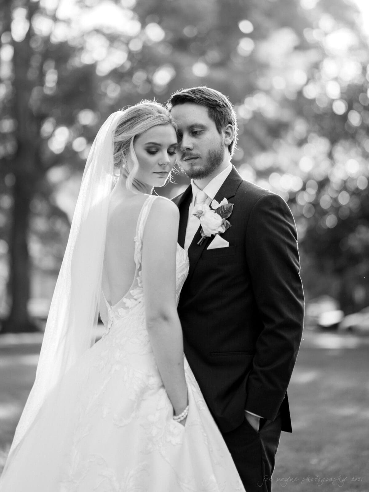 A bride and groom stand close together outdoors in a serene, wooded setting. The bride, in a lace gown and veil, looks down gently, while the groom, in a dark suit with a boutonniere, gazes at her. The scene is softly lit, emphasizing their calm expressions.