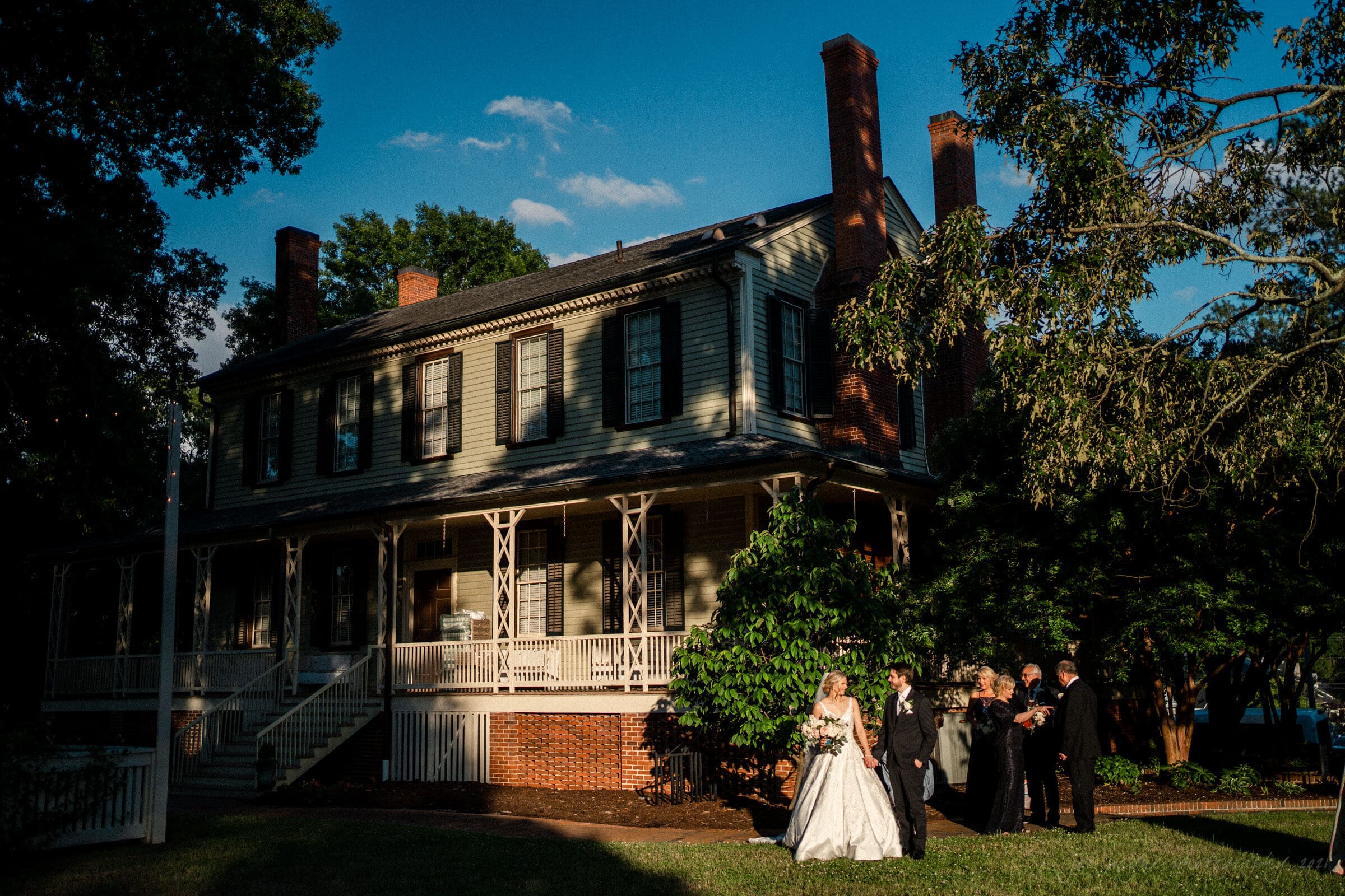 A bride and groom stand with a group of people in formal attire outside a large, historic house. The setting sun casts long shadows, highlighting the greenery and architecture. The sky is clear with a few clouds.