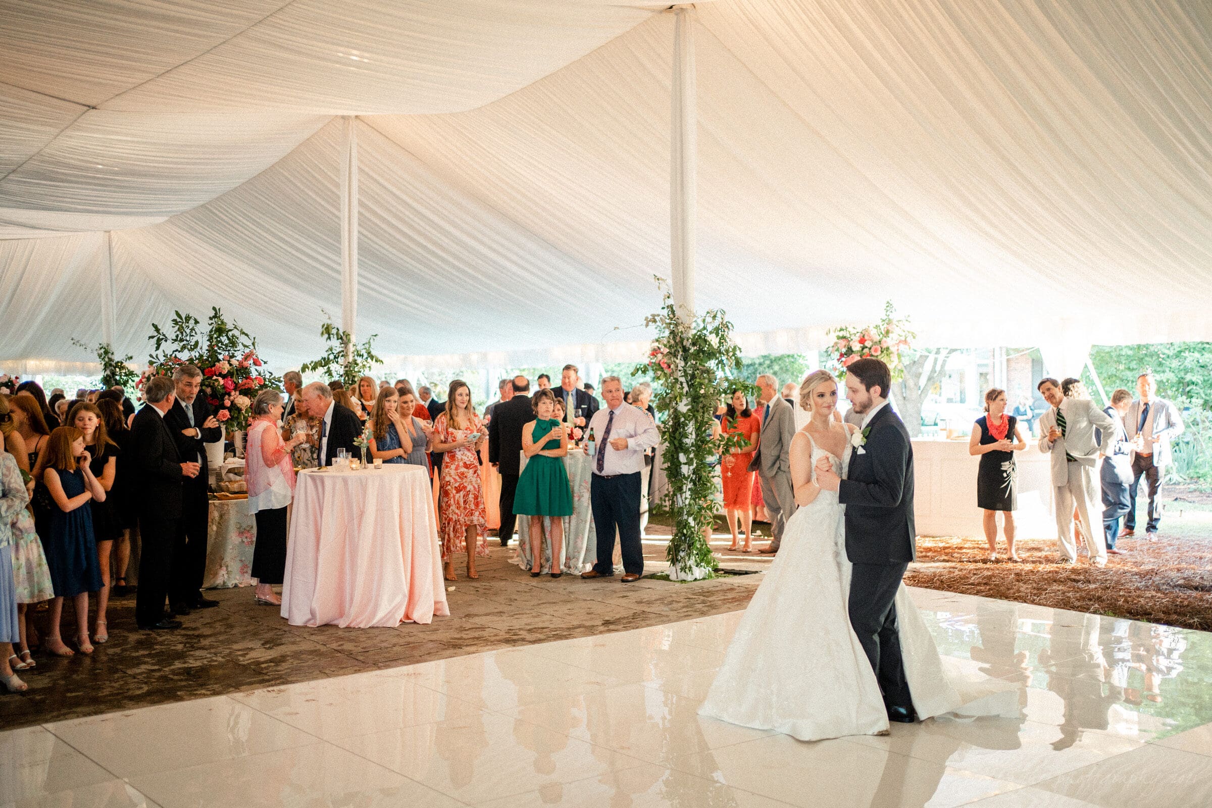 A bride and groom dance under a large white tent at a wedding reception, surrounded by elegantly dressed guests. Tables with floral arrangements and a bar are visible in the background, creating a festive atmosphere.