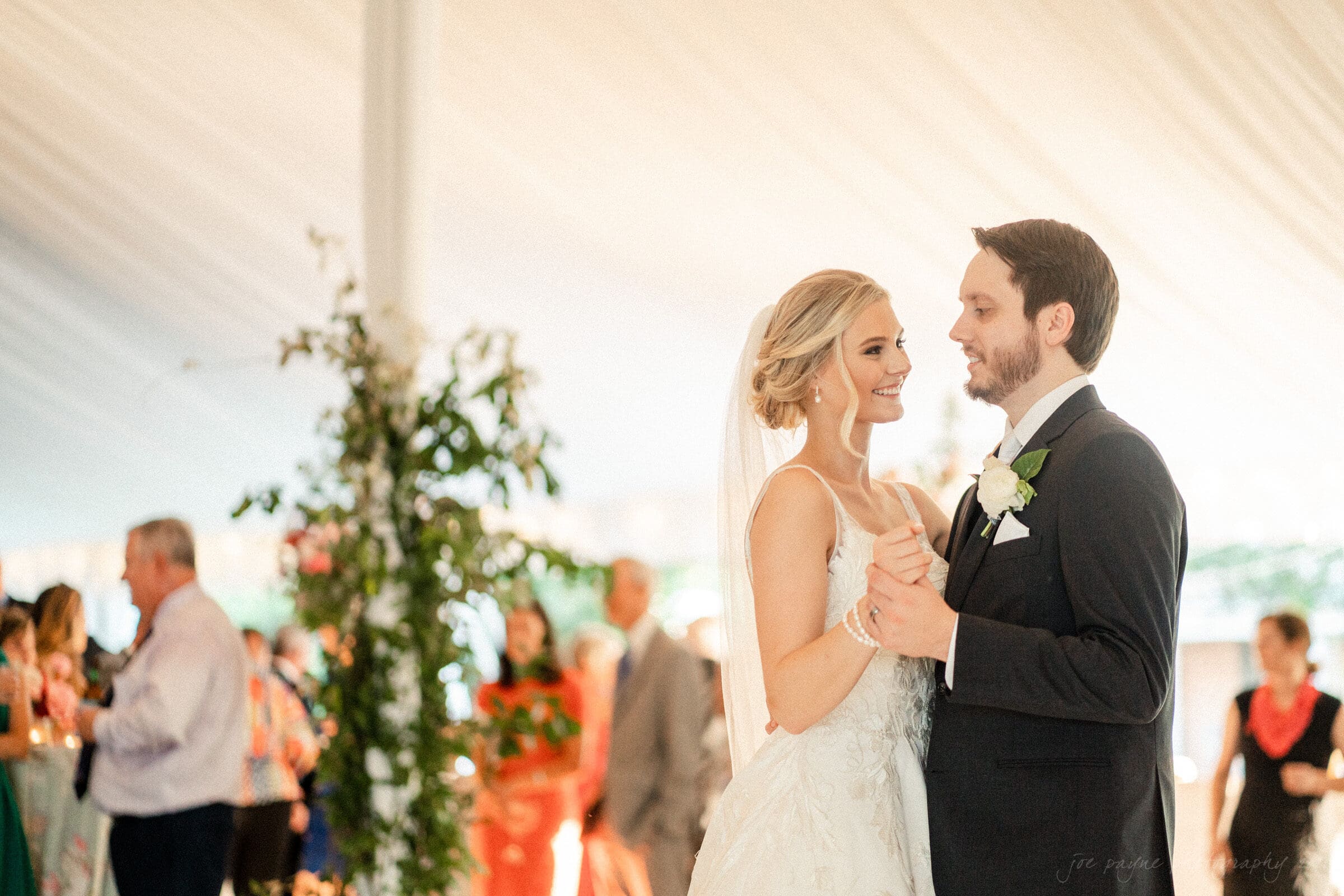 A bride and groom share their first dance at a wedding reception. Theyre smiling at each other under a decorated canopy, surrounded by softly blurred guests and greenery.