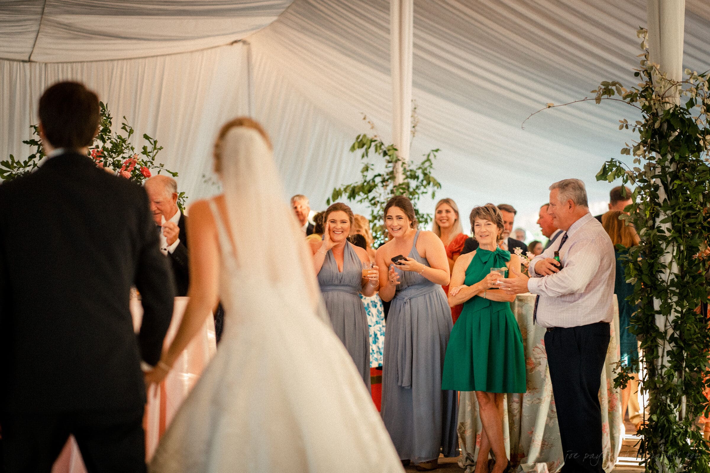 A bride and groom are walking towards a group of smiling guests at a wedding reception. The guests are standing under a white tent, dressed in formal attire, and are surrounded by lush greenery and floral decorations.