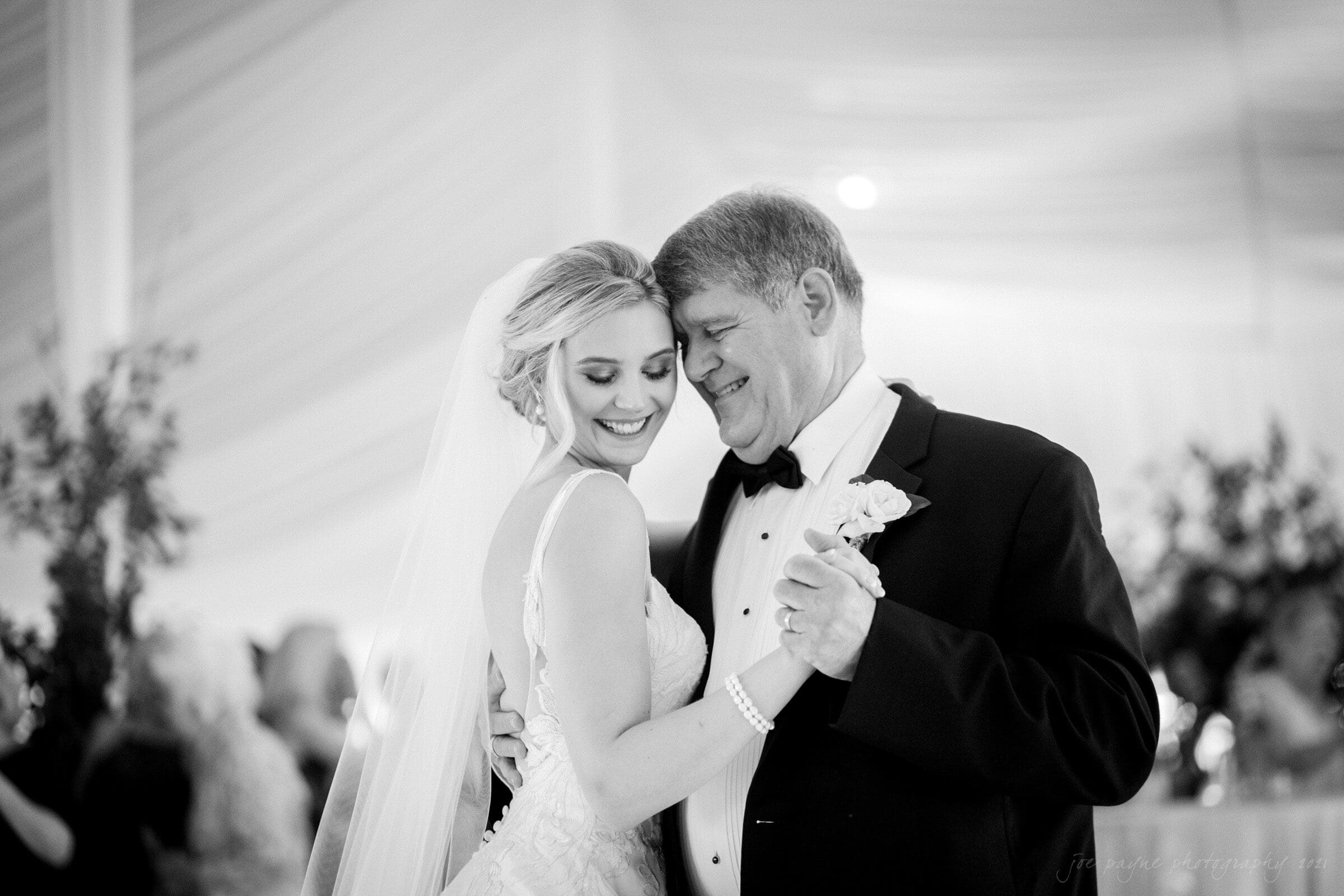 A bride in a wedding dress smiles while dancing with an older man in a tuxedo. They are in a warmly lit setting with a draped tent ceiling and blurred greenery in the background. Both appear joyful and close.