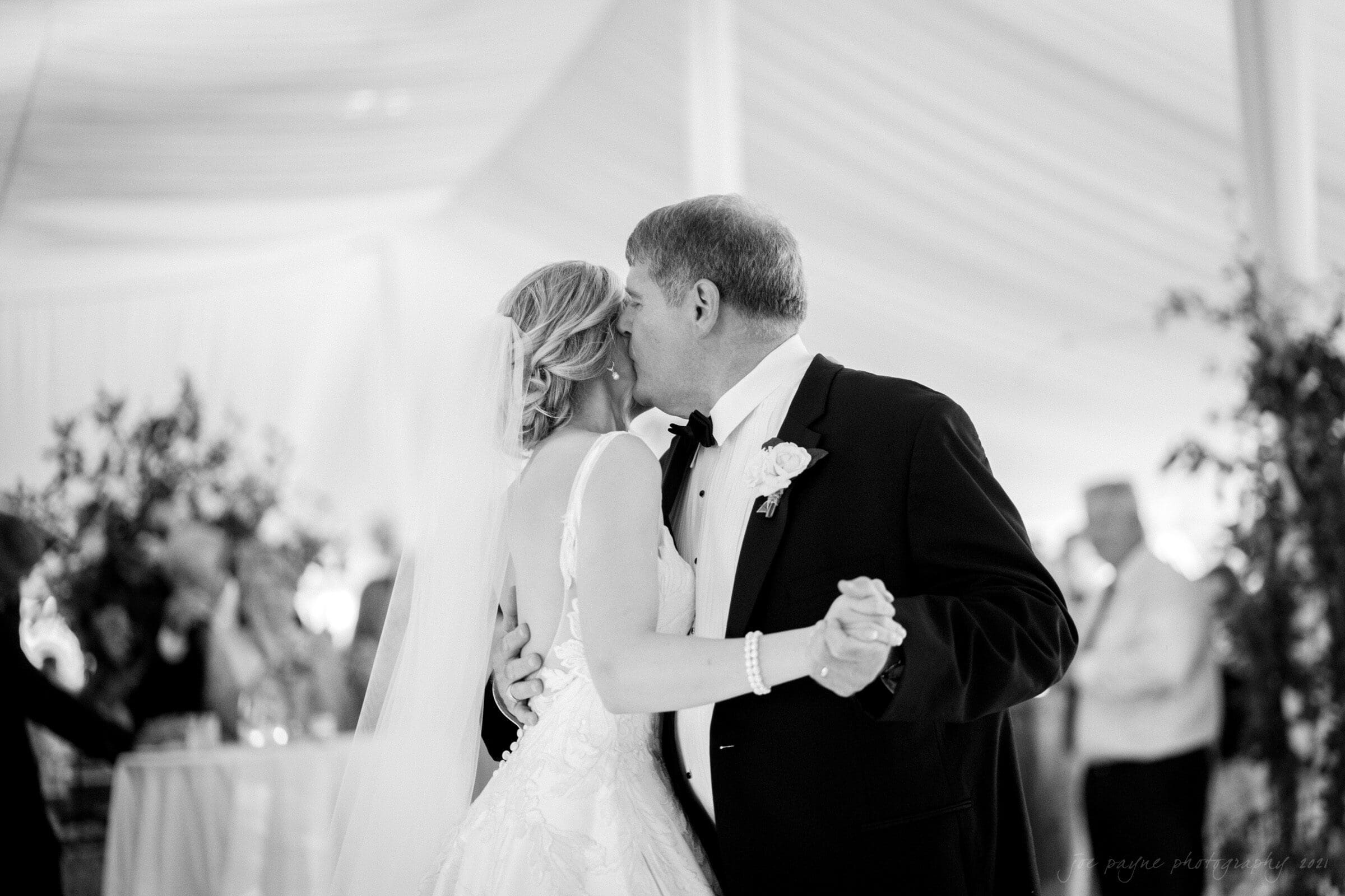 A black and white photo of a groom in a tuxedo dancing closely with his bride in a wedding dress. They are in a decorated venue with guests and plants in the background. The brides veil is visible, and they appear joyful.