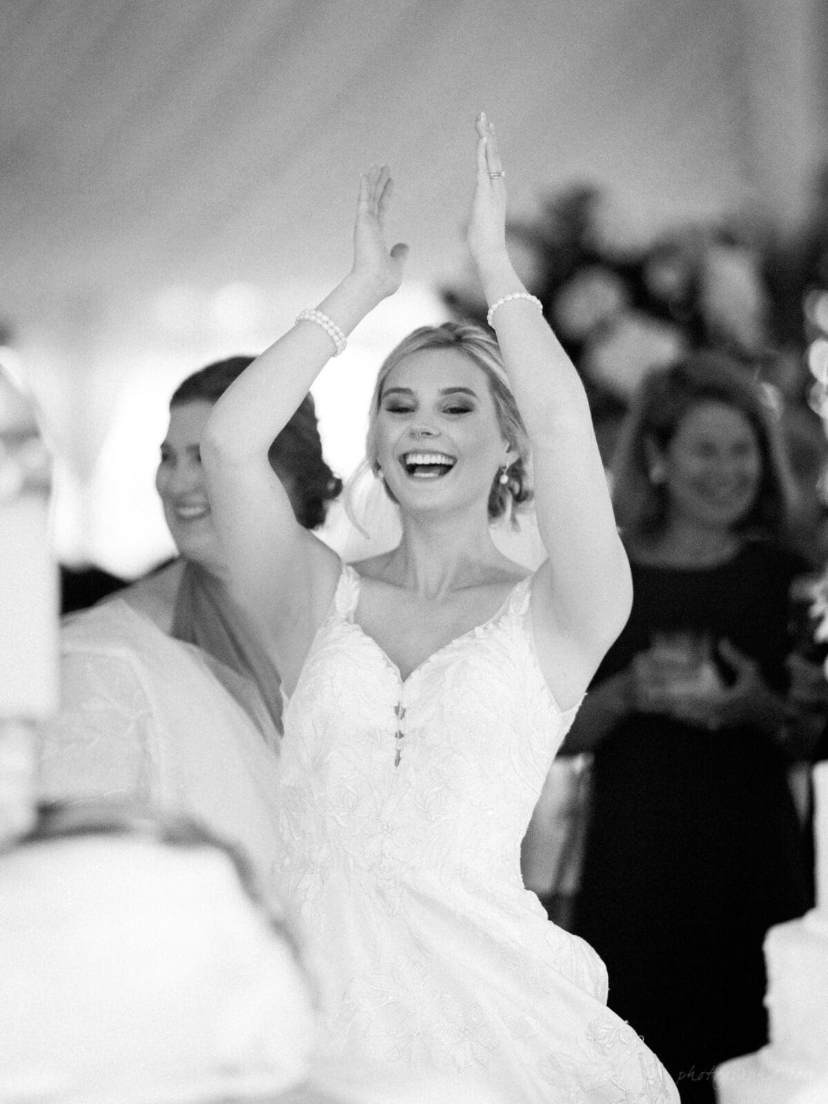 A joyful bride in a white dress is clapping and smiling widely at a wedding reception. She is surrounded by friends and family, with a blurred background that includes flowers and festive decor. The image is in black and white.