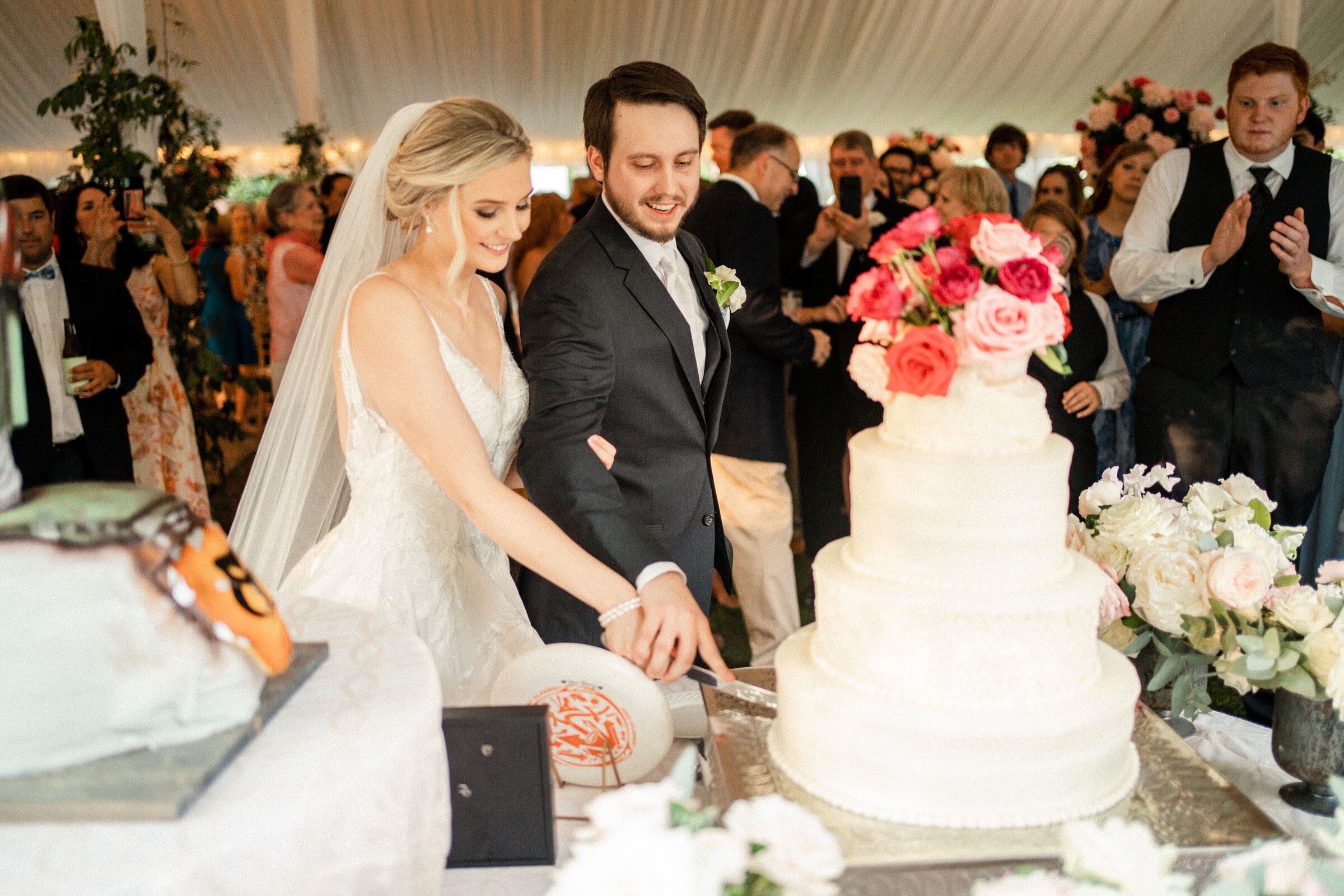 A bride and groom cut a large tiered wedding cake adorned with pink and white flowers. They are under a white tent, surrounded by guests. A decorated football helmet is visible in the foreground.
