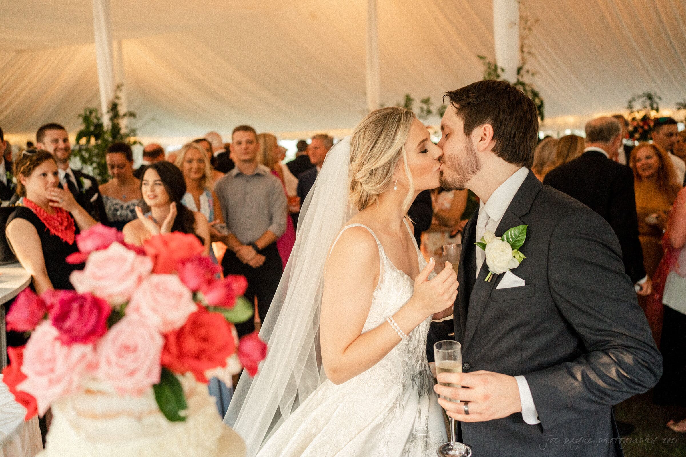 A bride and groom kiss under a white tent surrounded by guests. The bride wears a white dress and a veil, while the groom wears a suit with a white rose. A floral arrangement and part of a wedding cake are visible in the foreground.