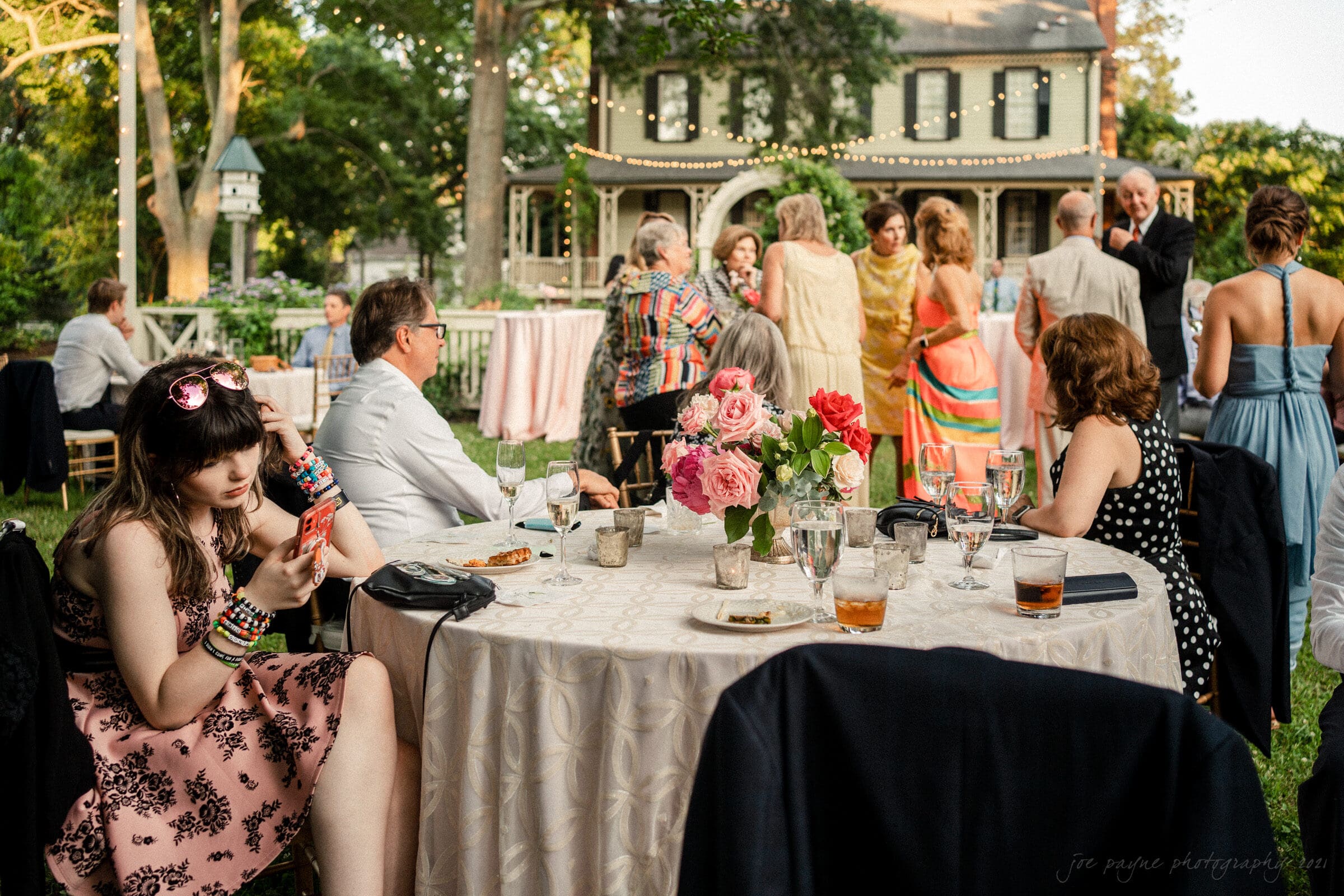 A young woman in a floral dress sits at a table with her phone, surrounded by empty glasses and plates. People in colorful attire dance and socialize in front of a house adorned with string lights in the background.
