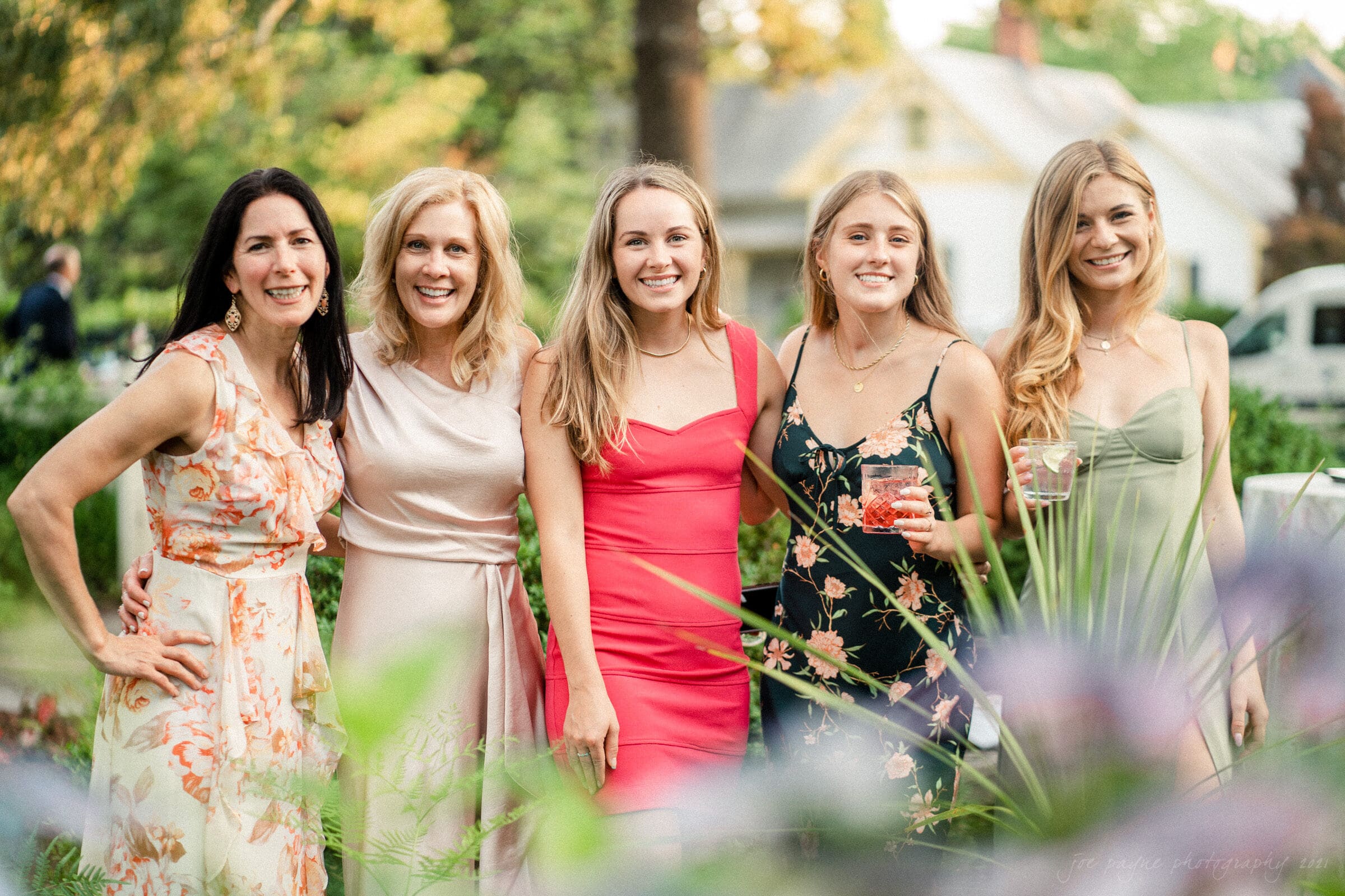 Five women are standing together outdoors, smiling at the camera. They are dressed in colorful summer dresses, surrounded by greenery, with a house visible in the background. Some hold drinks in their hands.