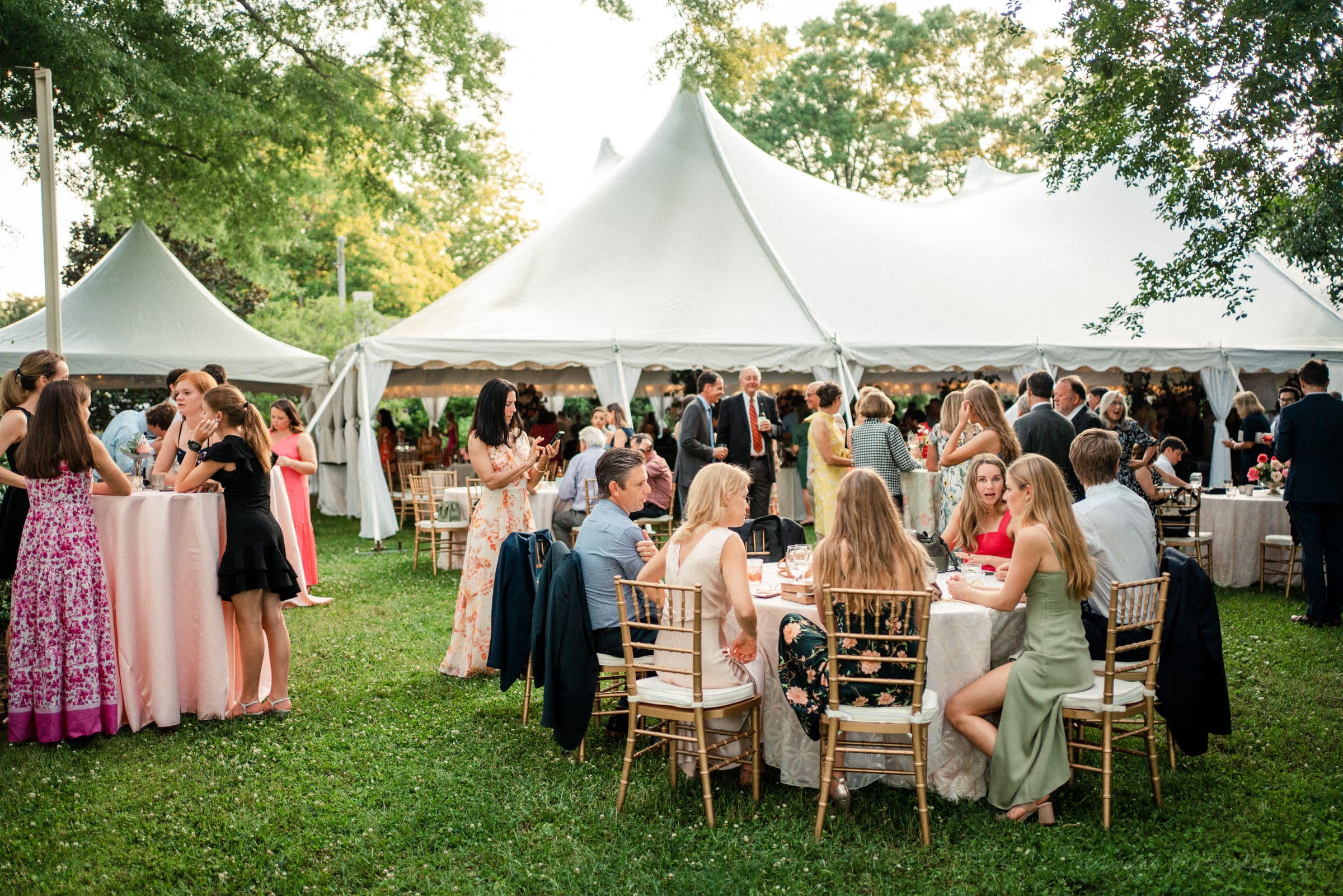A lively outdoor gathering under large white tents, with elegantly dressed guests seated at round tables on a grassy lawn. People are socializing and enjoying the event in a vibrant, green setting.