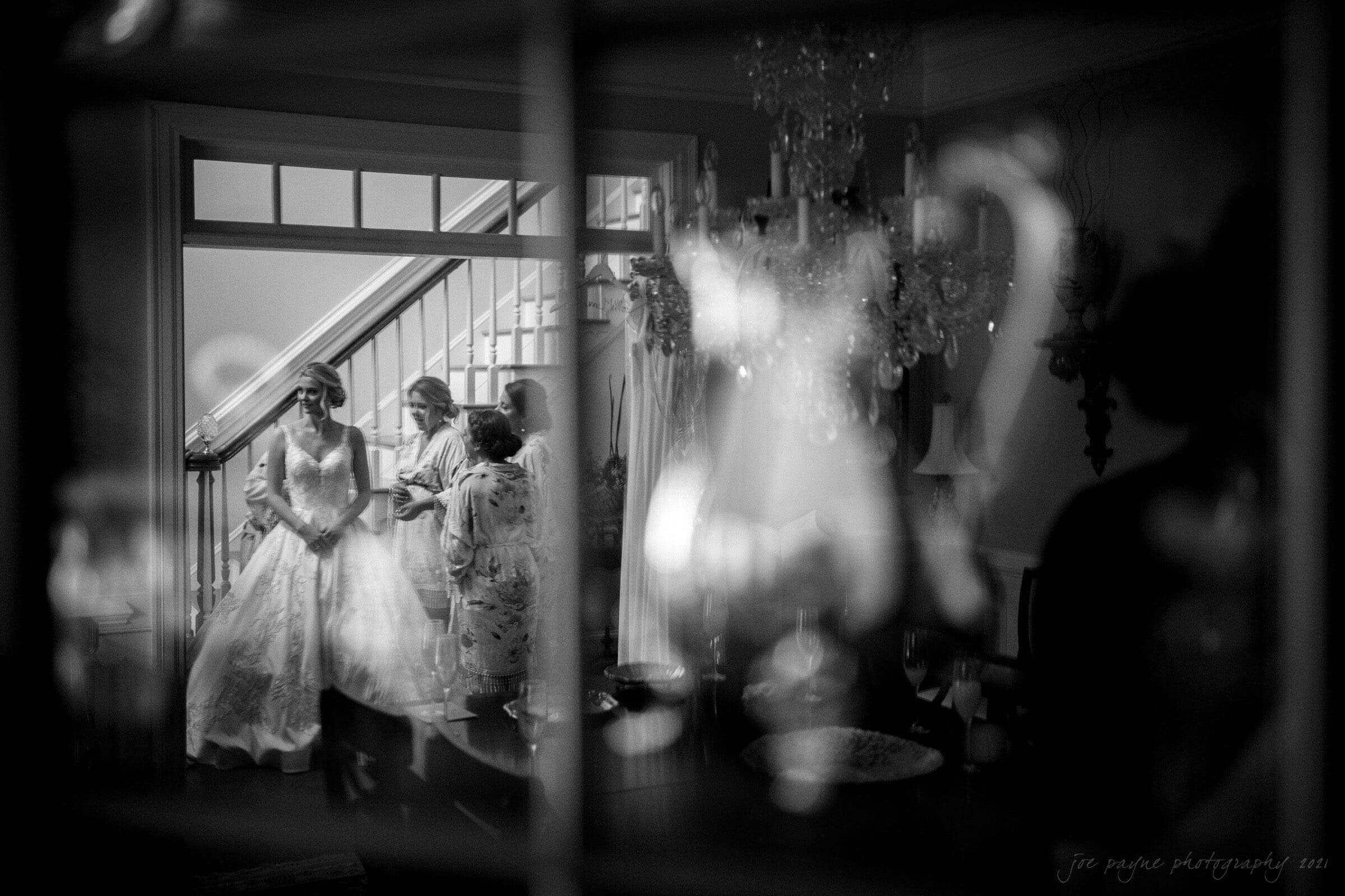 Black and white image of a bride in a gown standing by a window with two women. They are indoors, with a chandelier in the foreground. The scene is viewed through a reflective surface, adding a dreamy effect.