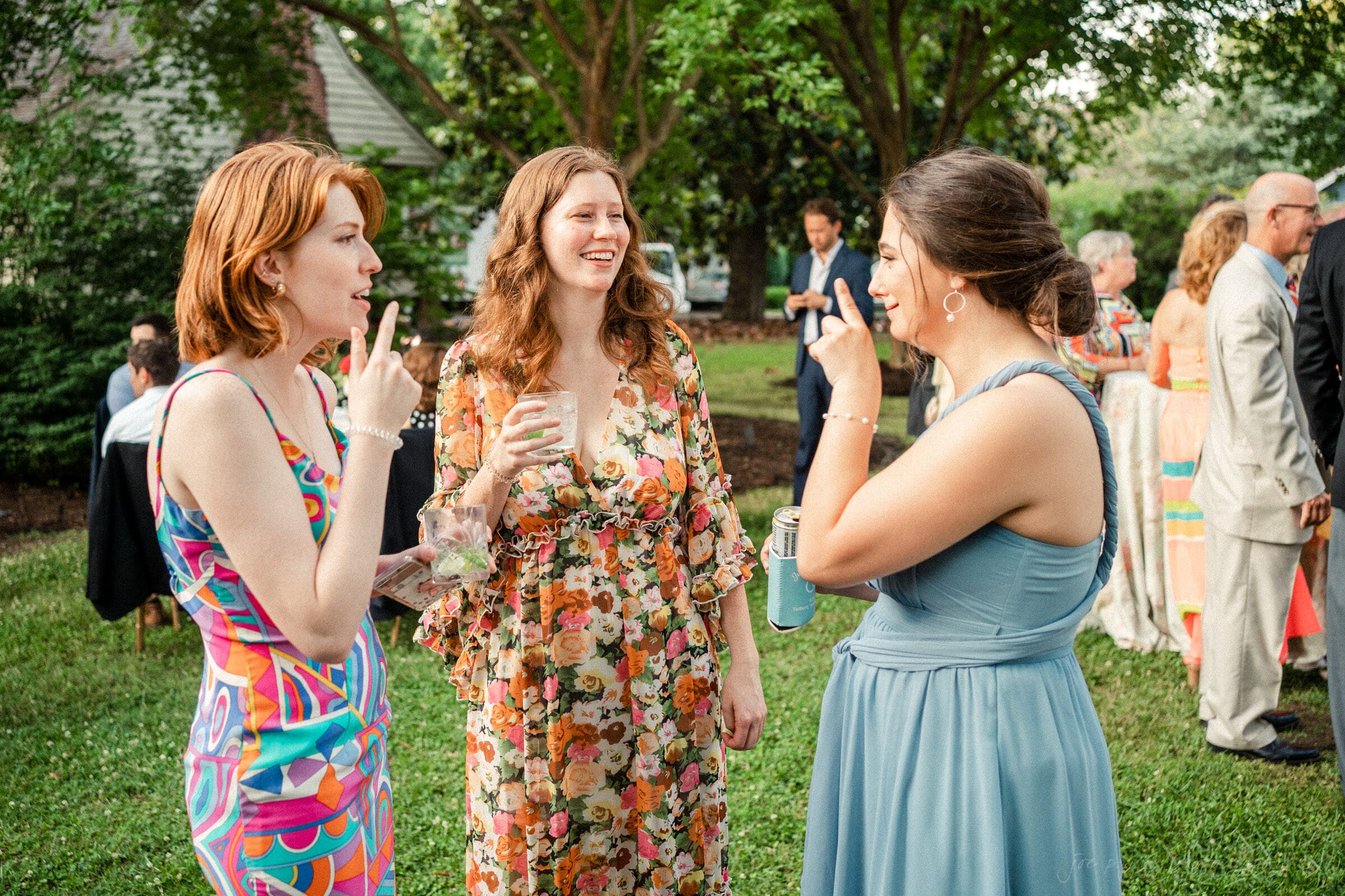 Three women are standing and smiling while holding drinks at an outdoor gathering. They are dressed in colorful summer dresses. In the background, other guests are mingling amidst greenery and trees.