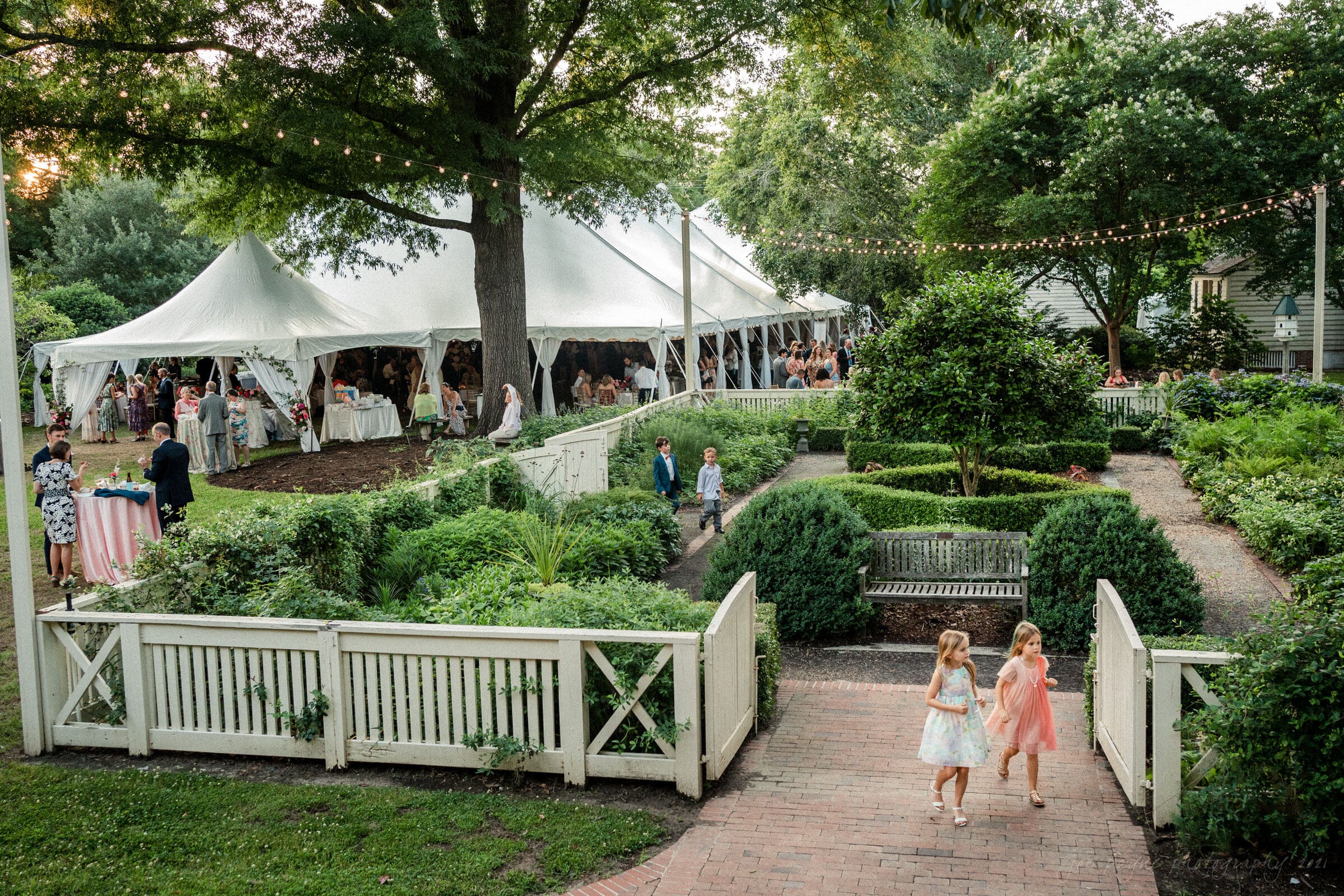 A garden wedding reception with a white tent, surrounded by lush greenery and fairy lights. Guests gather under the tent, while two children walk along a brick path in the foreground, bordered by a white fence and neatly trimmed bushes.