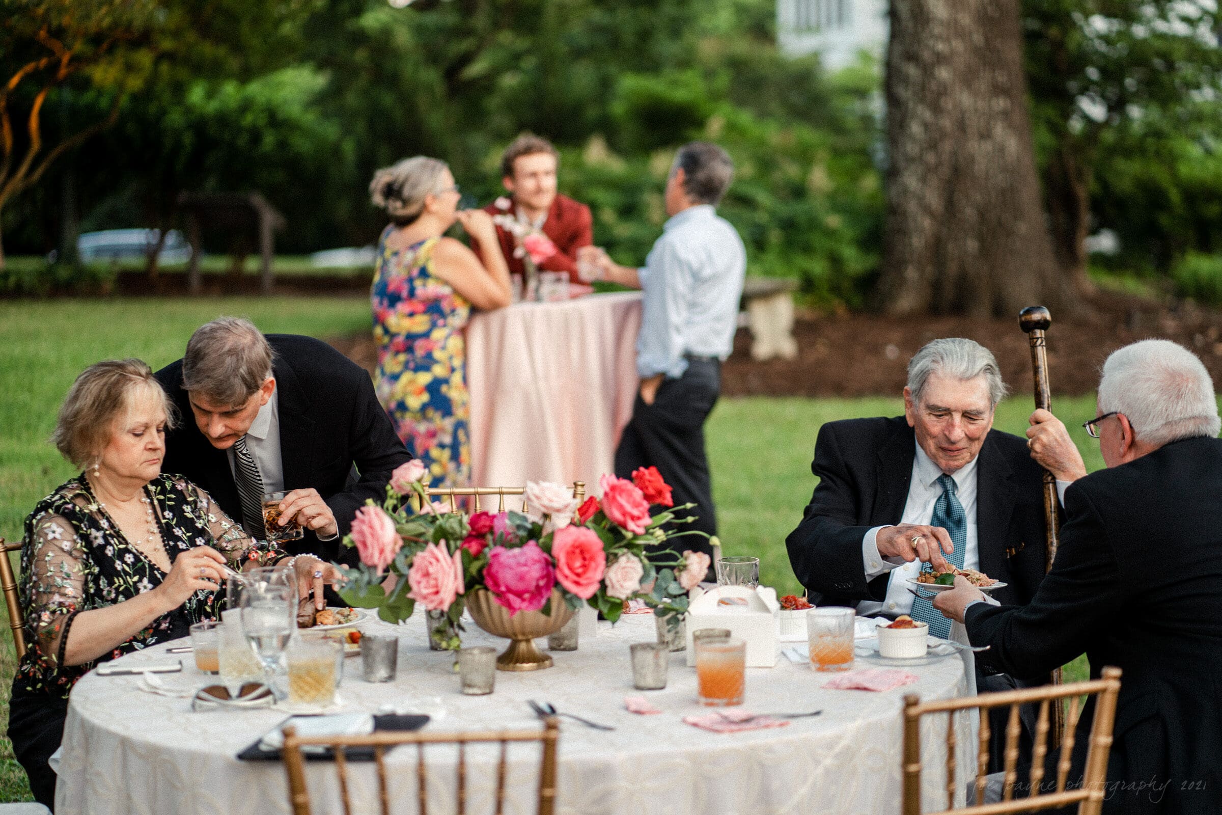A group of people at an outdoor event, with some seated around a table decorated with flowers and others standing and chatting in the background. The scene is set in a garden with trees and lush greenery.