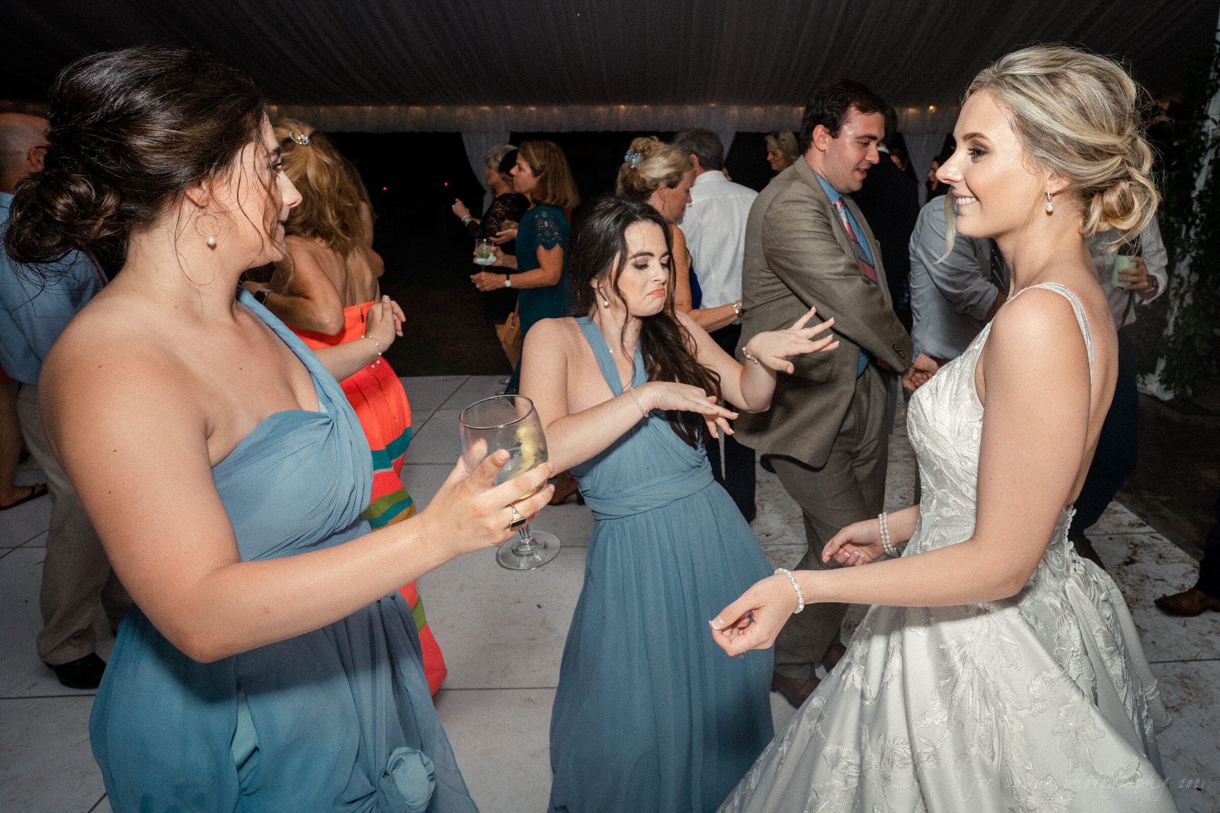 Three women in formal dresses enjoy dancing at a party. One in a wedding gown smiles, while the others in blue dresses hold drinks. Guests chat in the background under a tent.