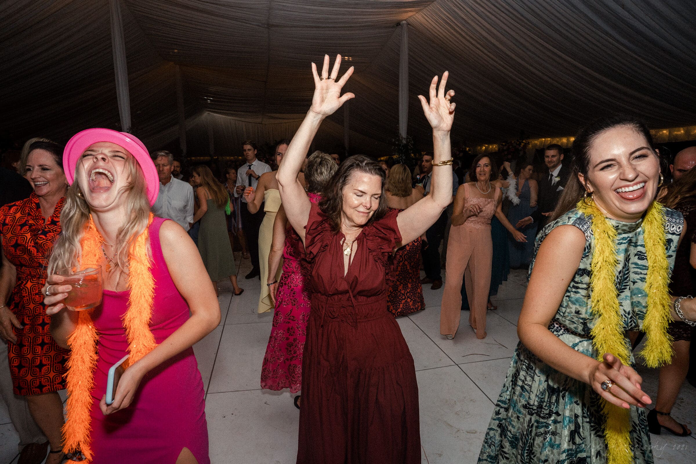 A lively indoor party scene with people dancing and enjoying themselves. One woman in a pink hat laughs with a drink, while another raises her hands joyfully. The setting is under a large tent with dim lighting. Colorful outfits and decorations add vibrancy.