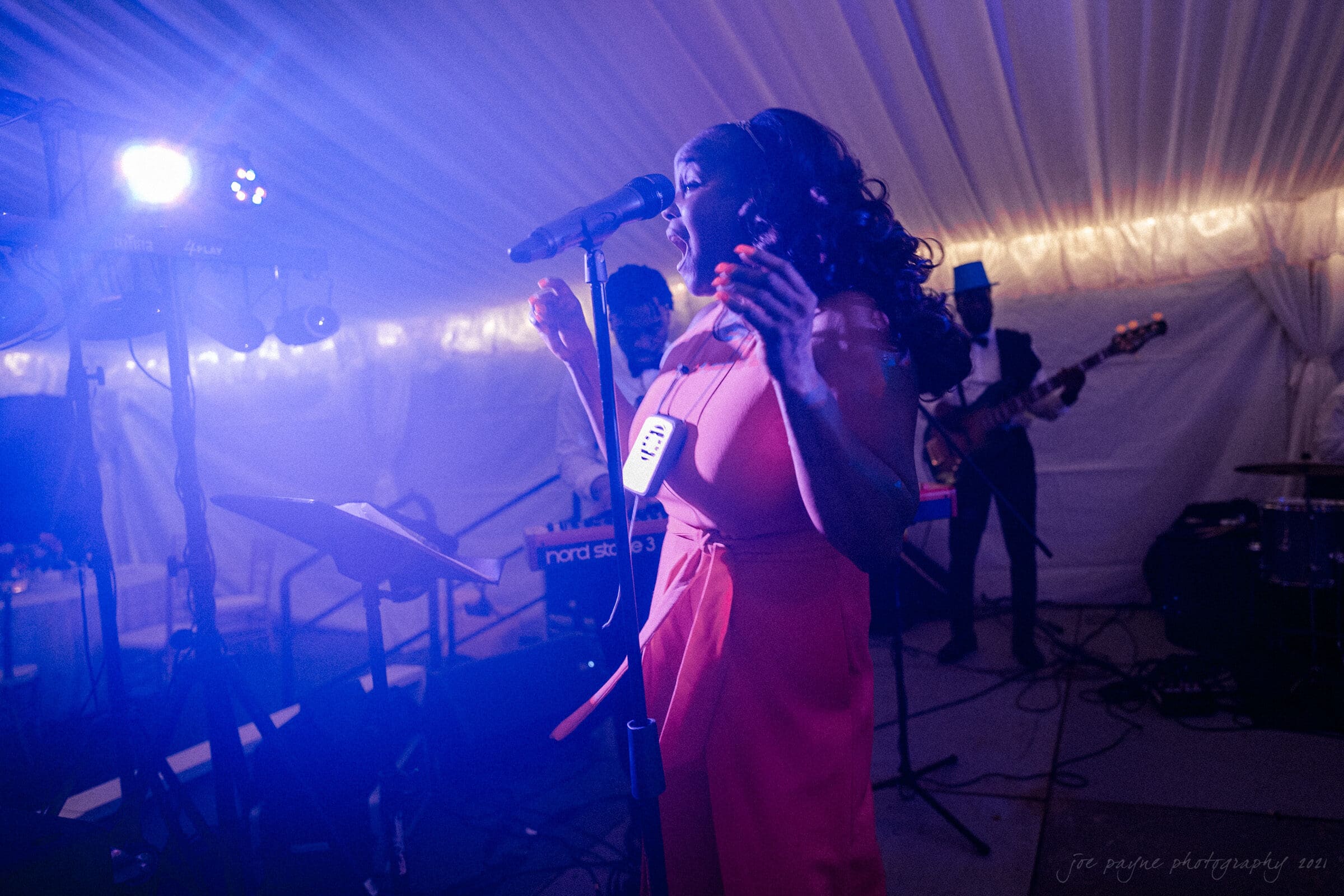 A woman in a pink dress sings passionately into a microphone under soft stage lighting, accompanied by a band in the background, inside a tent decorated with string lights.