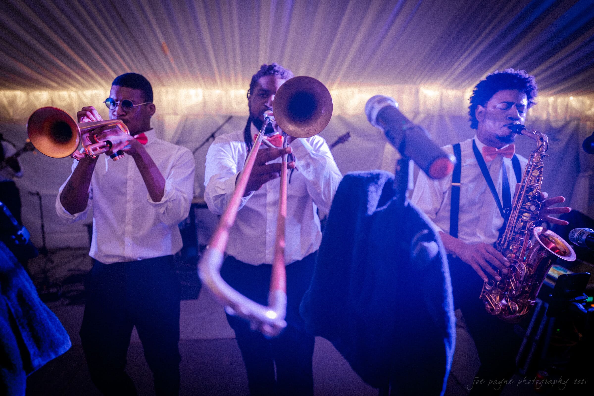 Three musicians perform energetically at an indoor event. A trumpet player, trombone player, and saxophonist wear white shirts and red bow ties, illuminated by soft stage lighting. A microphone stands in the foreground.