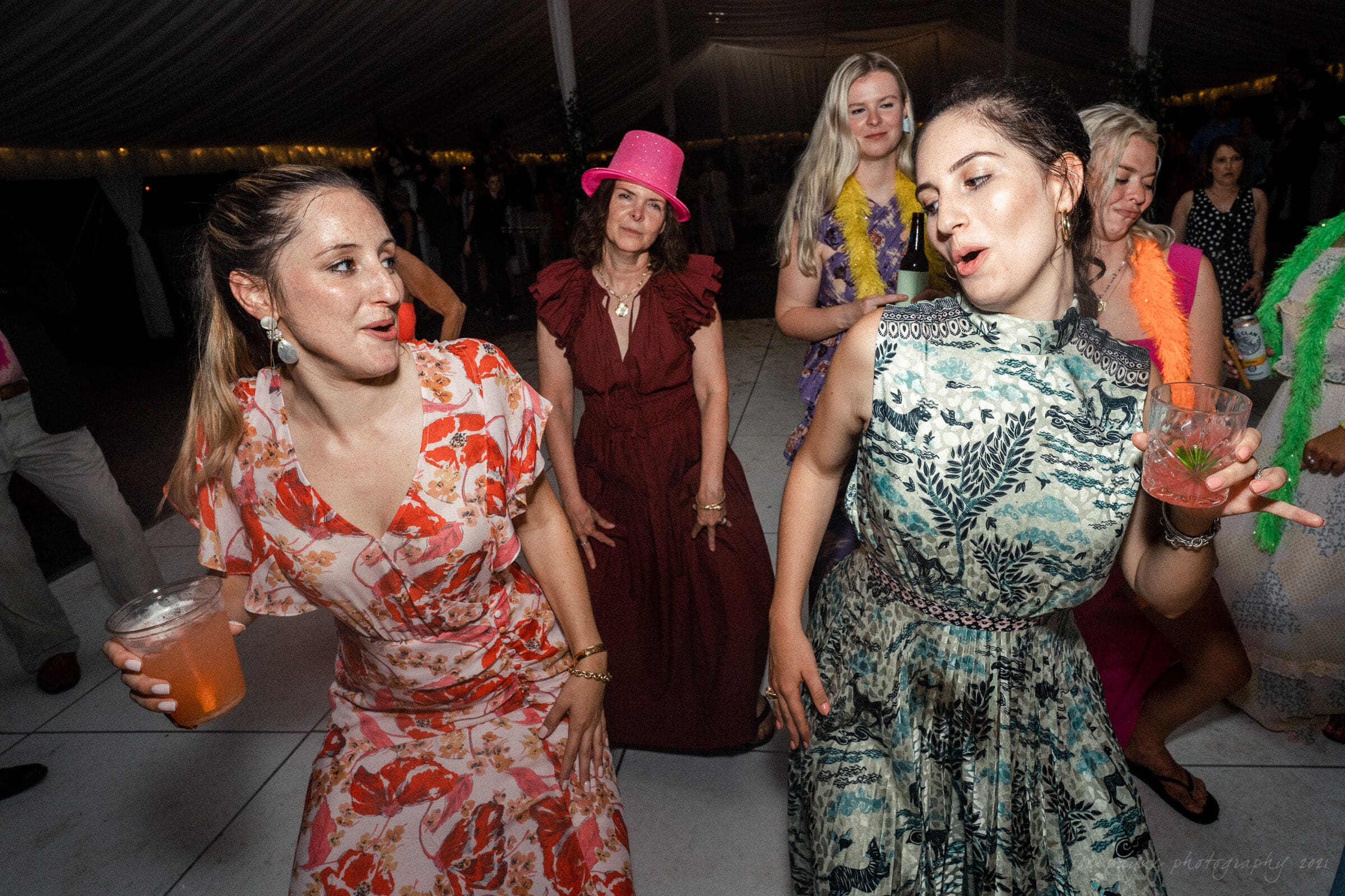 A group of people dancing energetically at an indoor event. Two women in the foreground are wearing floral dresses and holding drinks. A woman in the background wears a pink hat. The scene is lively and festive.